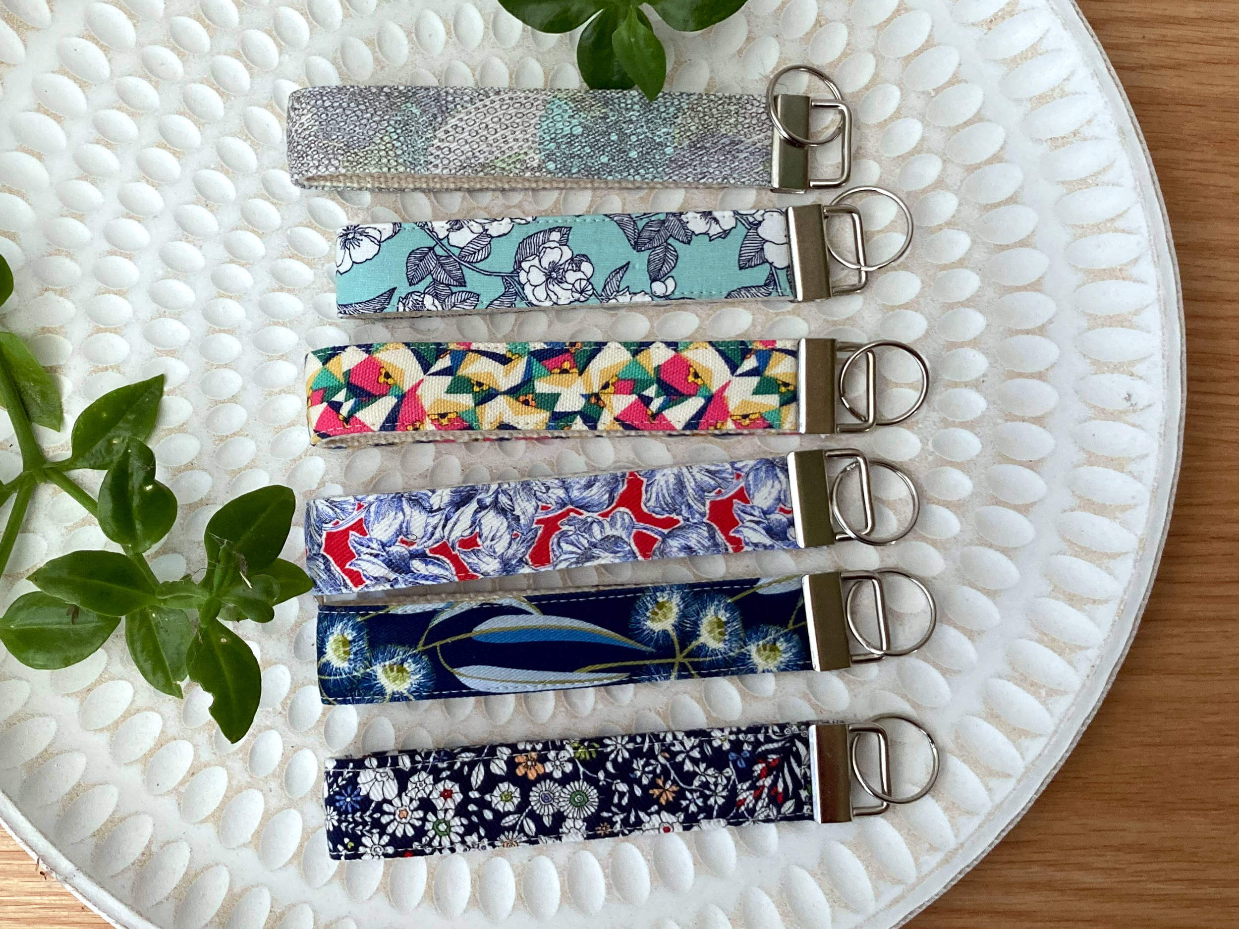 Three Key fob wristlets in various prints on a white textured plate with some plant greenery