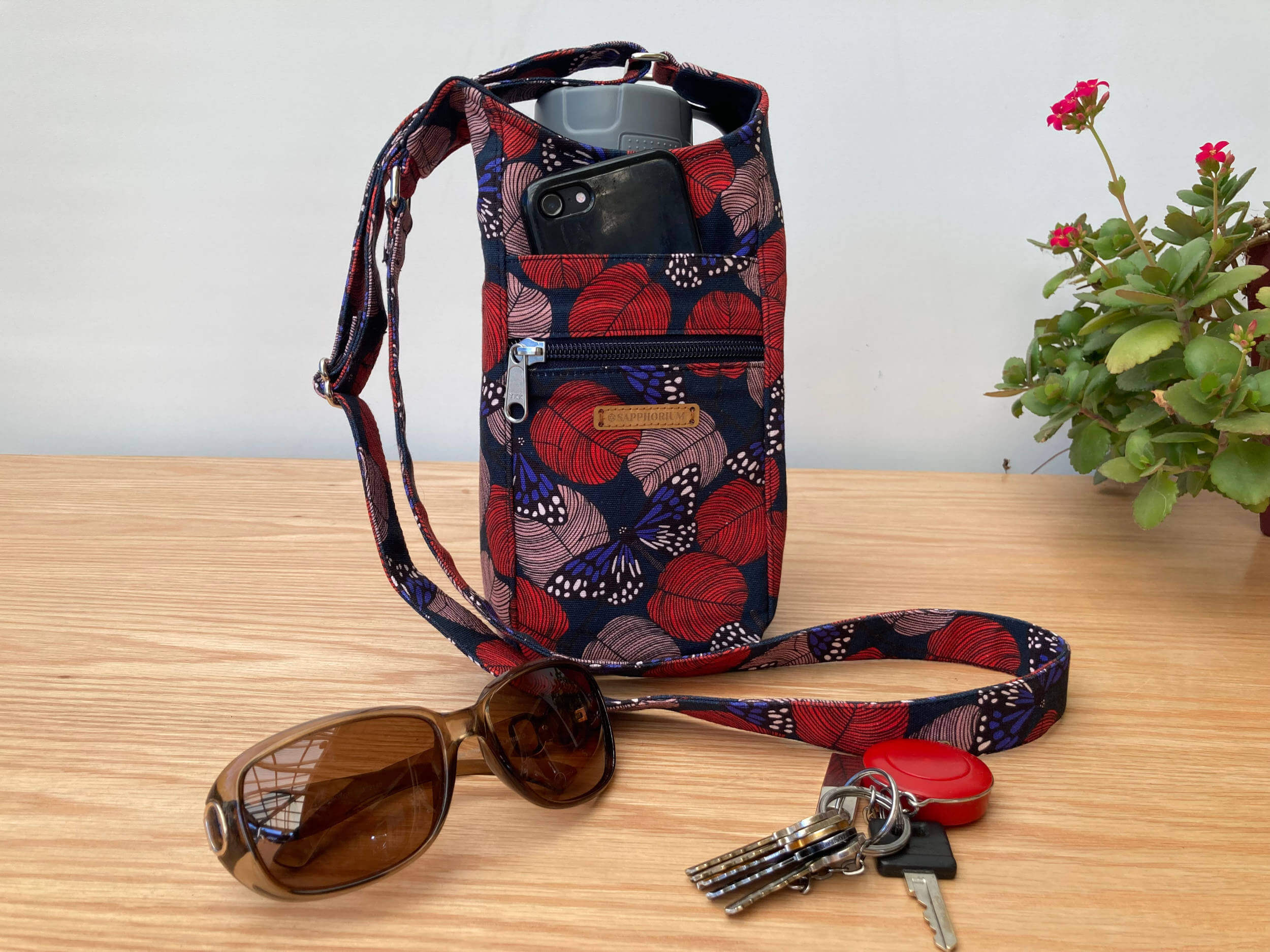 Navy Butterfly Water Bottle Bag by Sapphorium on table with sunglasses, keys, and plant