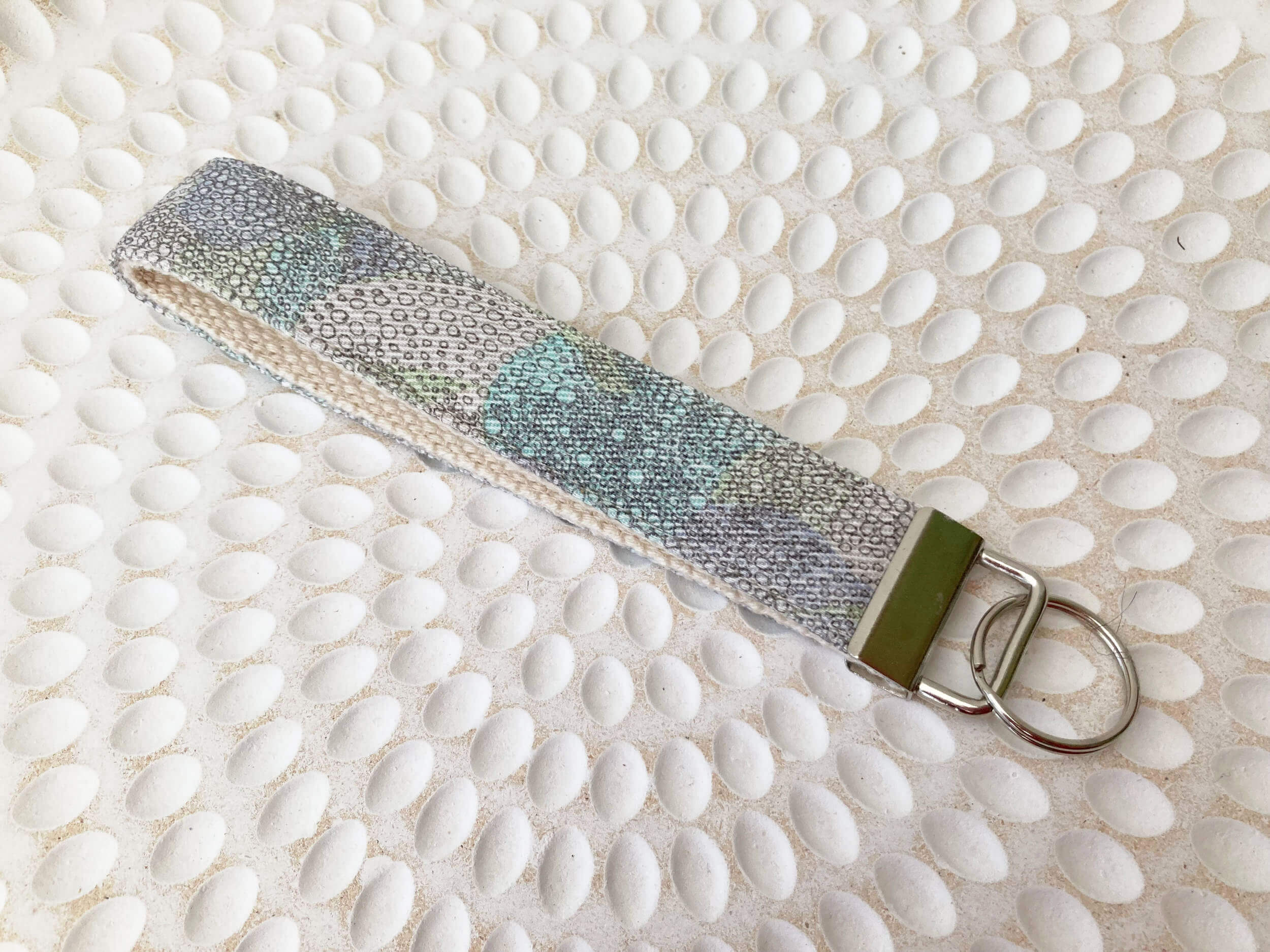 Key fob with small grey circles and various shades of pastel pink green on blue. Nickel Hardware. Laying on a textured plate.