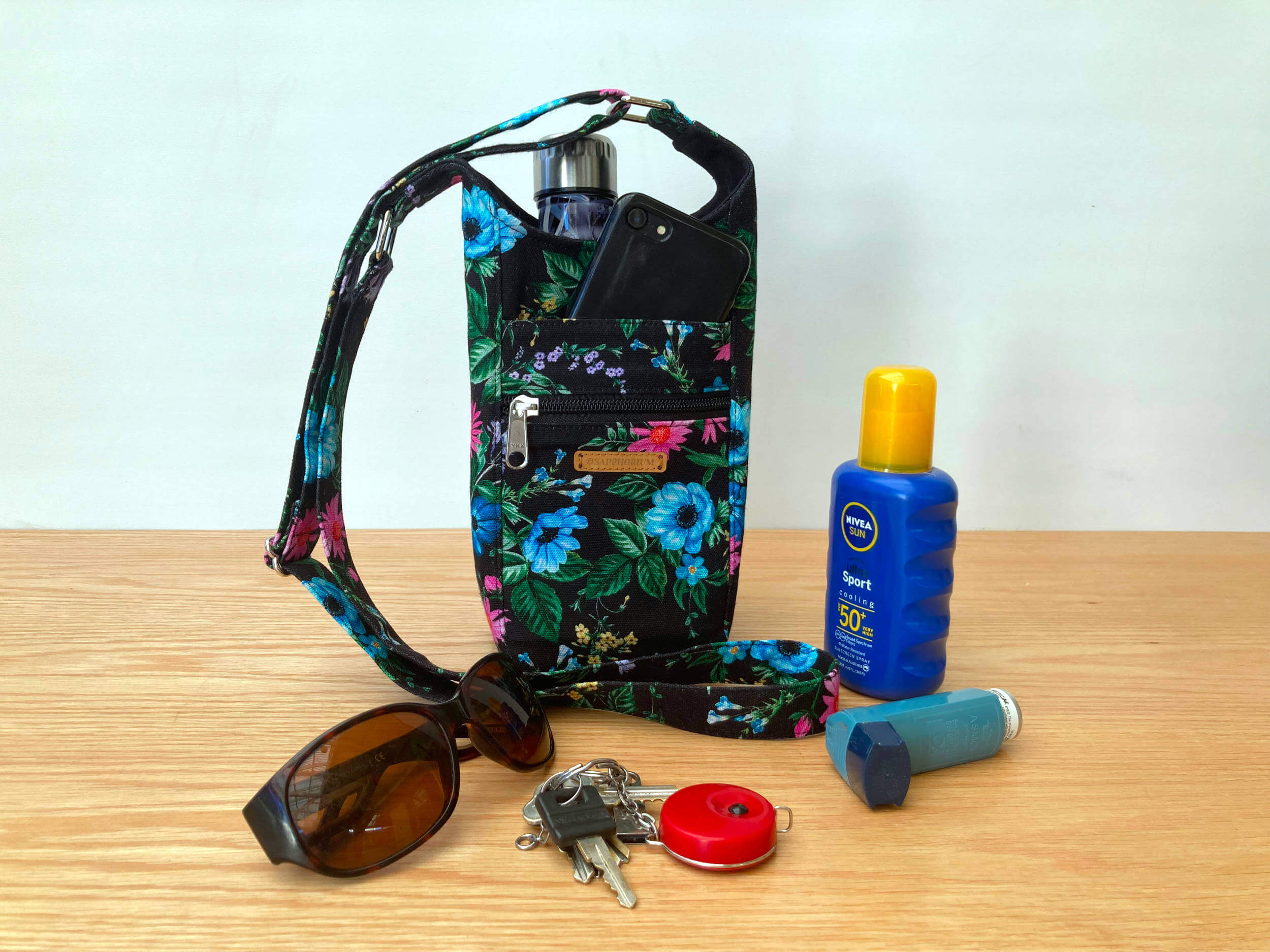 Black Floral Water Bottle Bag on table with water bottle inside it, and sunglasses, keys, asthma puffer and sunscreen bottle next to it.