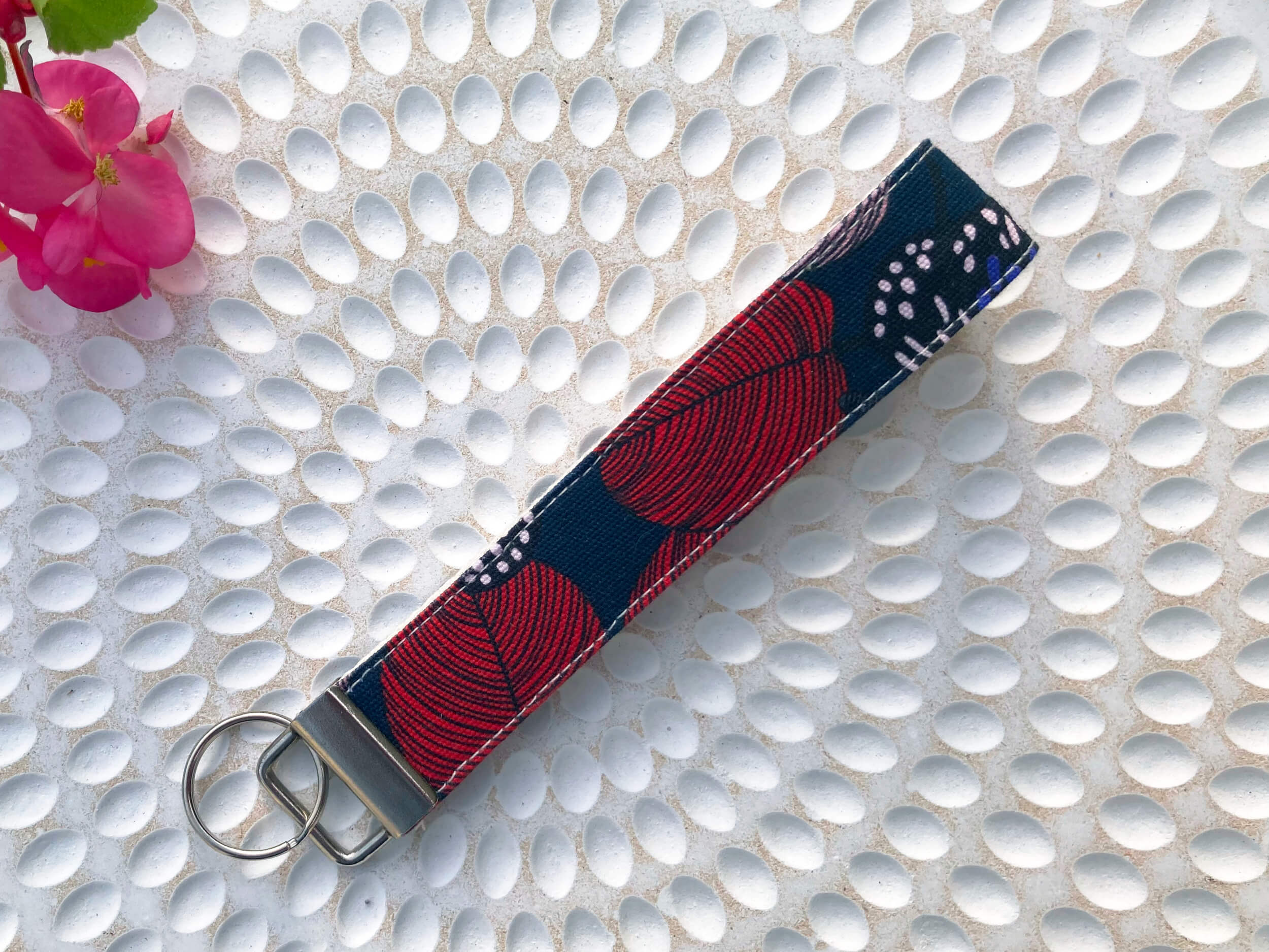 Key Fob with red leaves and part of a butterfly on navy background. Placed on textured background with pink flower and leaves in corner.