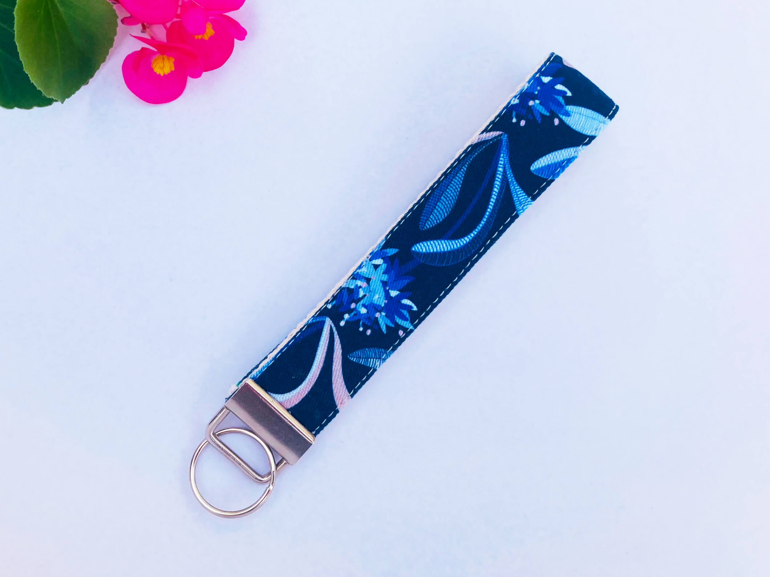 Fabric key fob with navy pincushion floral print by Jocelyn Proust. Laid on white background with pink flowers and green leaves in the corner.