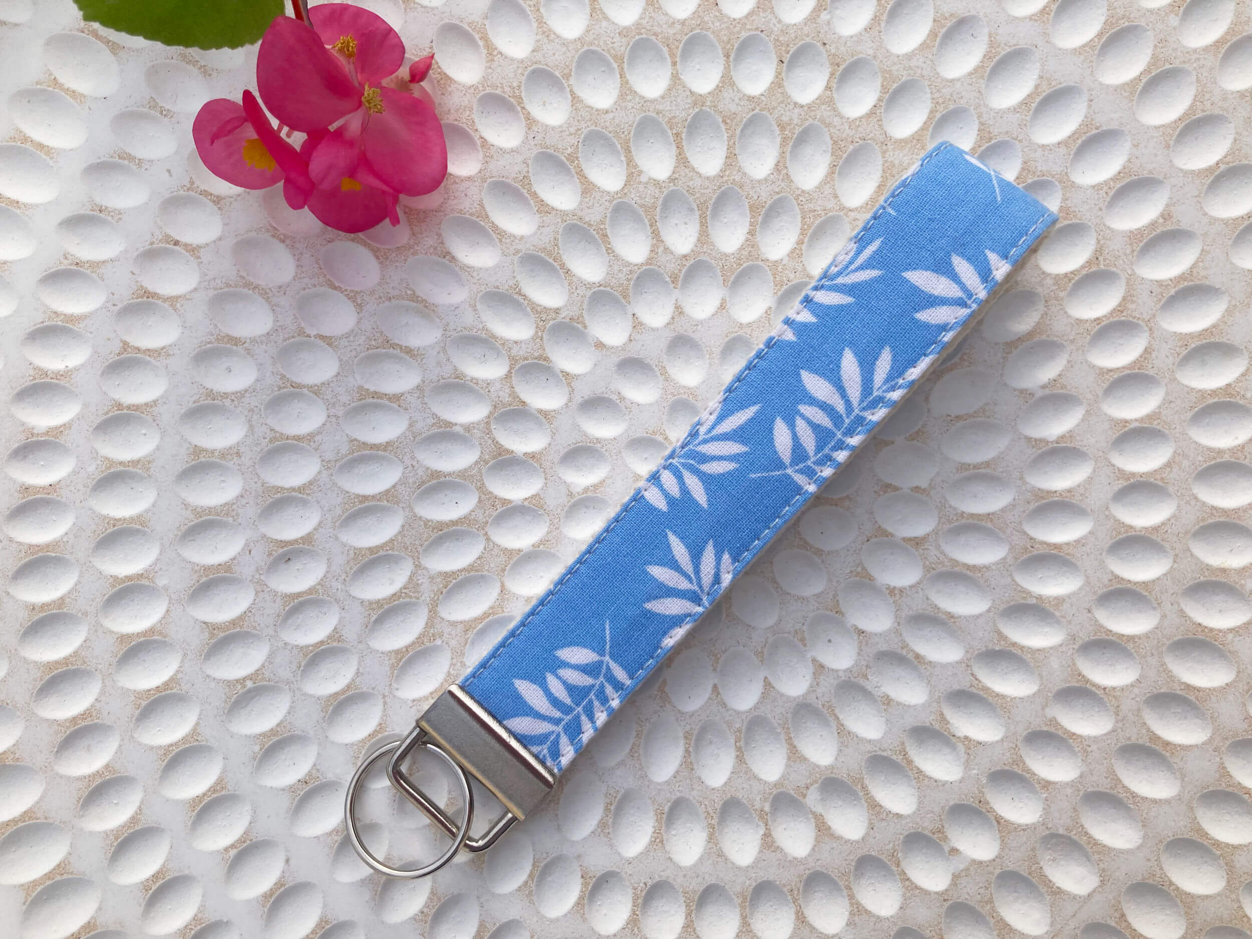 Key fob in a print with white leaves and light blue background. Item is on a textured background with pink flower in the corner.