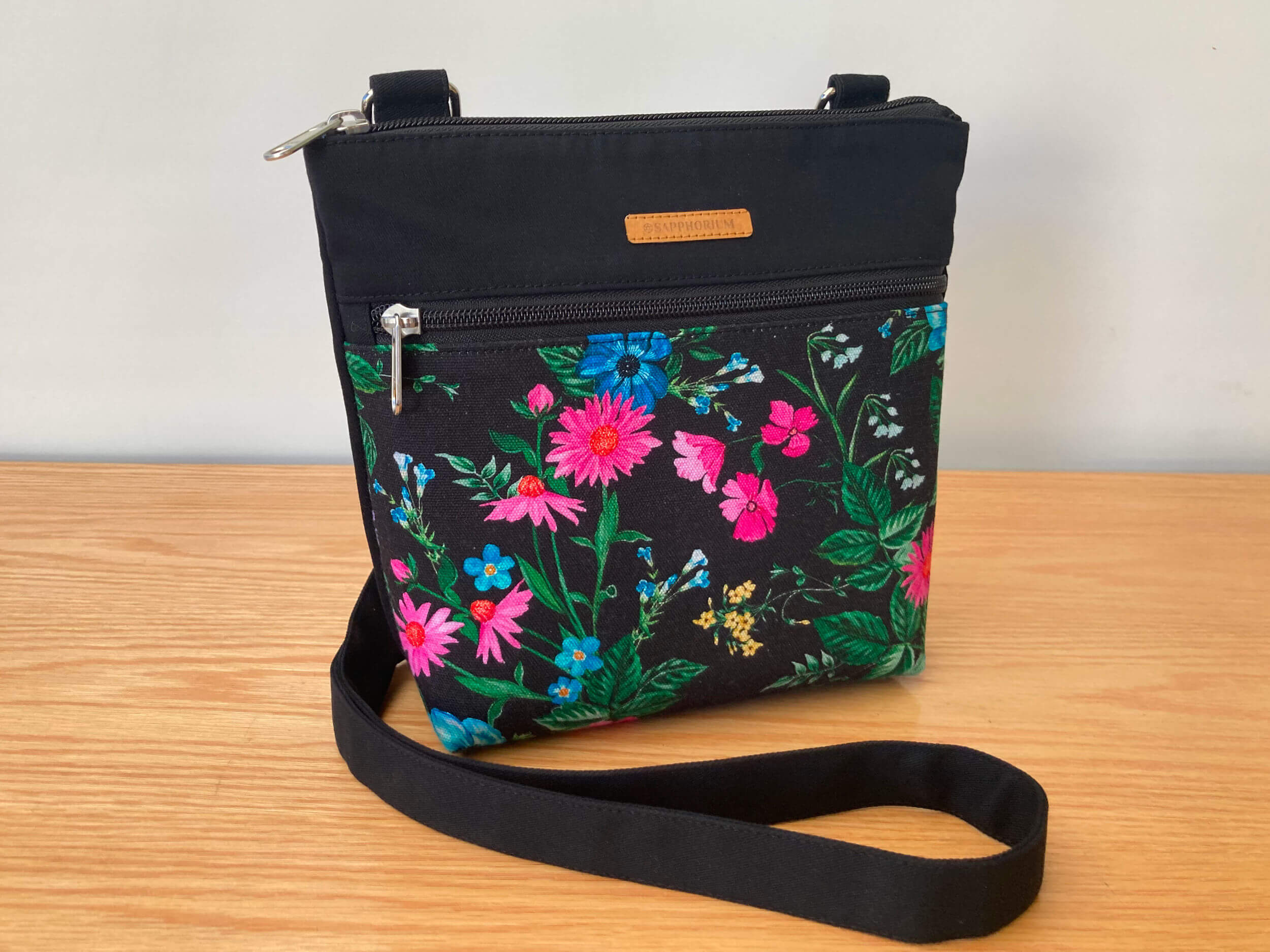 Black Crossbody bag with a floral print on a wooden table and white background.