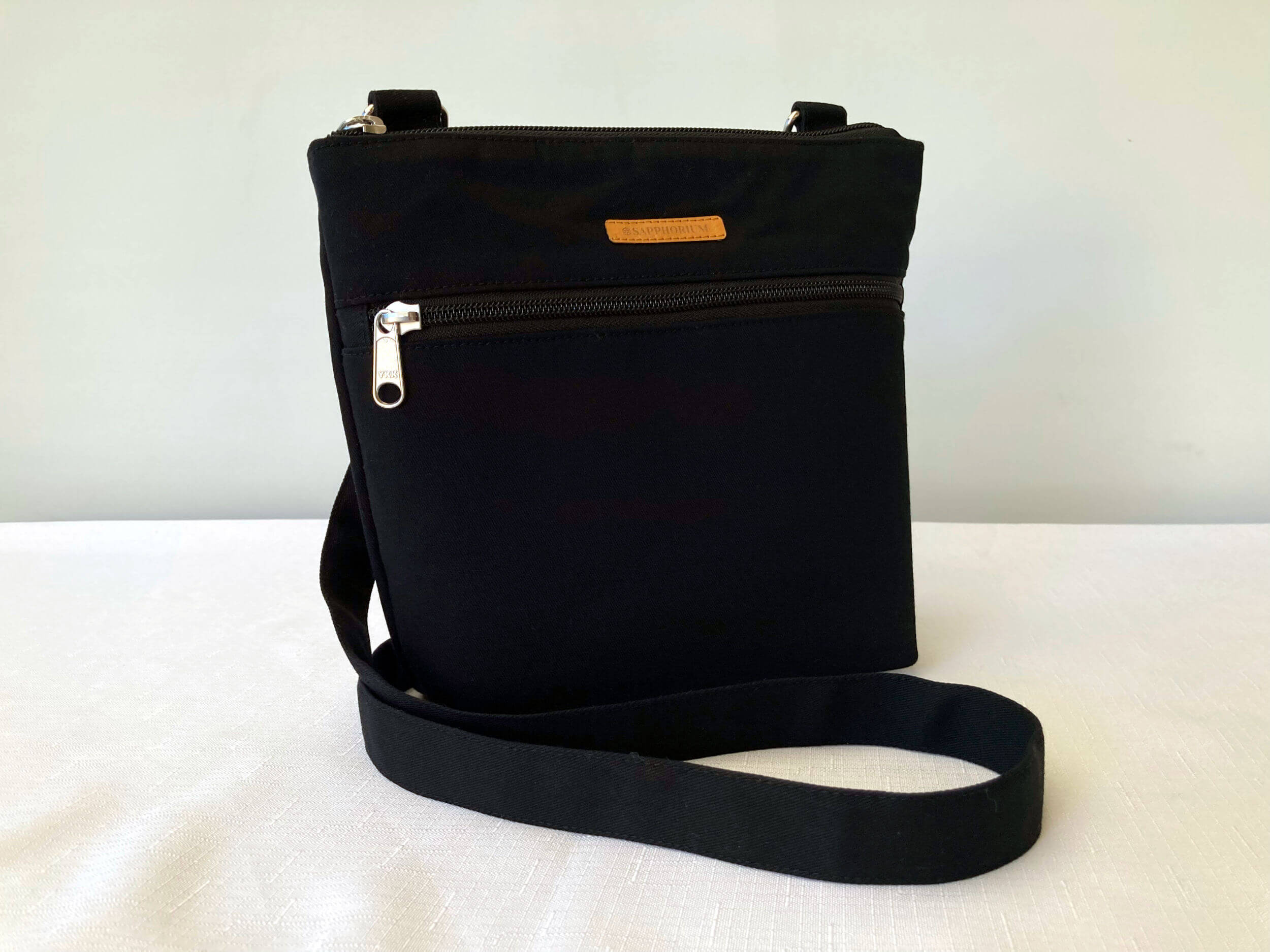 Black Crossbody bag with a top zip and front zip, both zipper pulls are silver. Brown label with Sapphorium. White background.