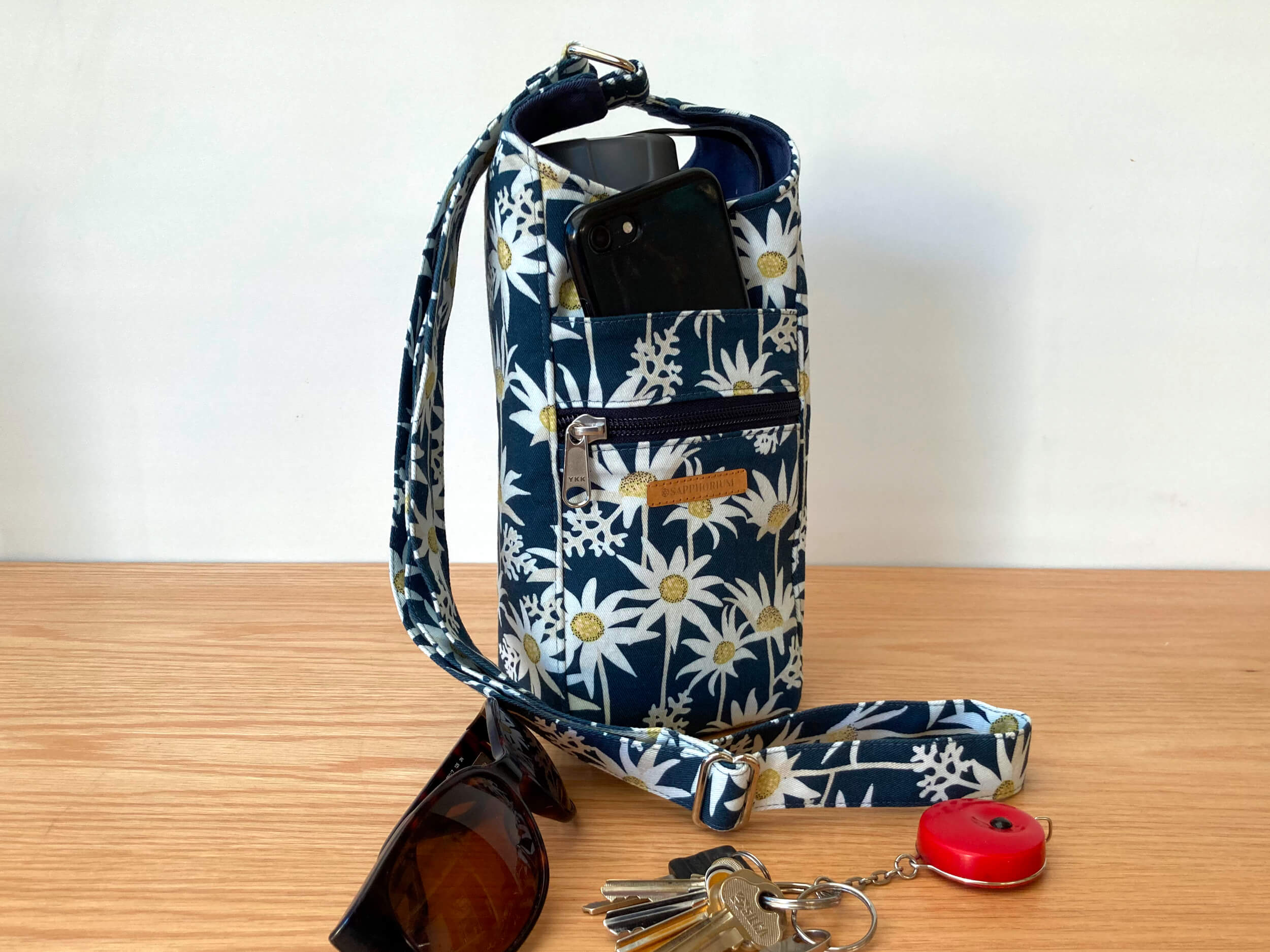 Water Bottle Bag with White Flannel Flowers on Marine Blue Print on a wooden table with sunglasses and keys.