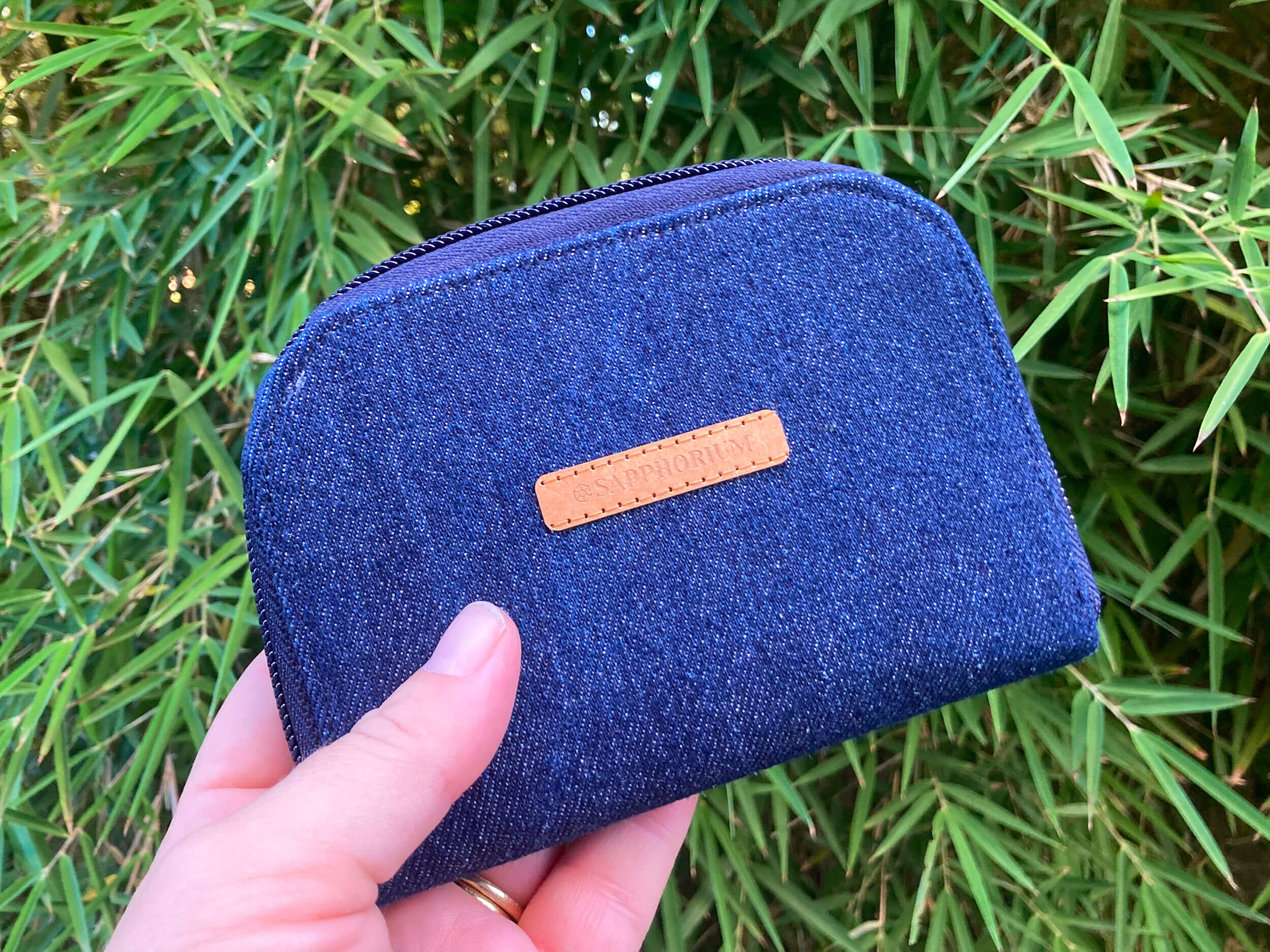 Denim zip wallet with brown label saying Sapphorium. Being held in front of leafy background