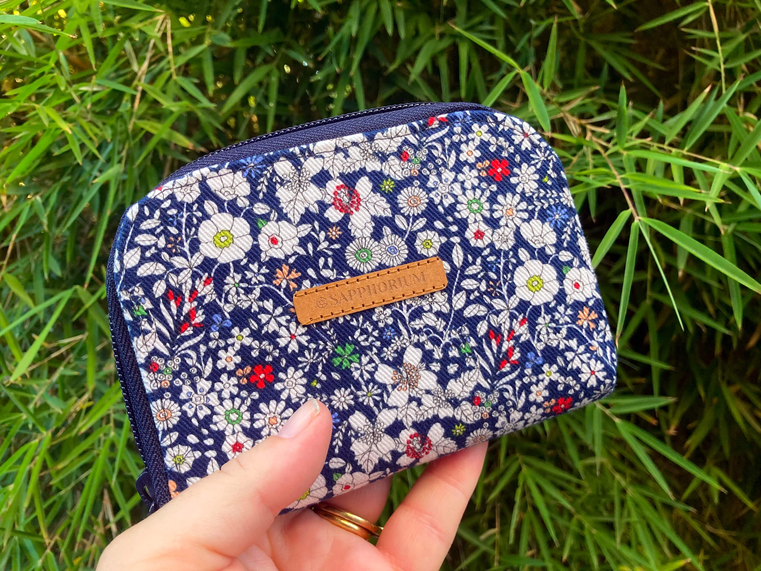 Blue and white floral zip wallet with brown label saying Sapphorium. Being held with leafy background