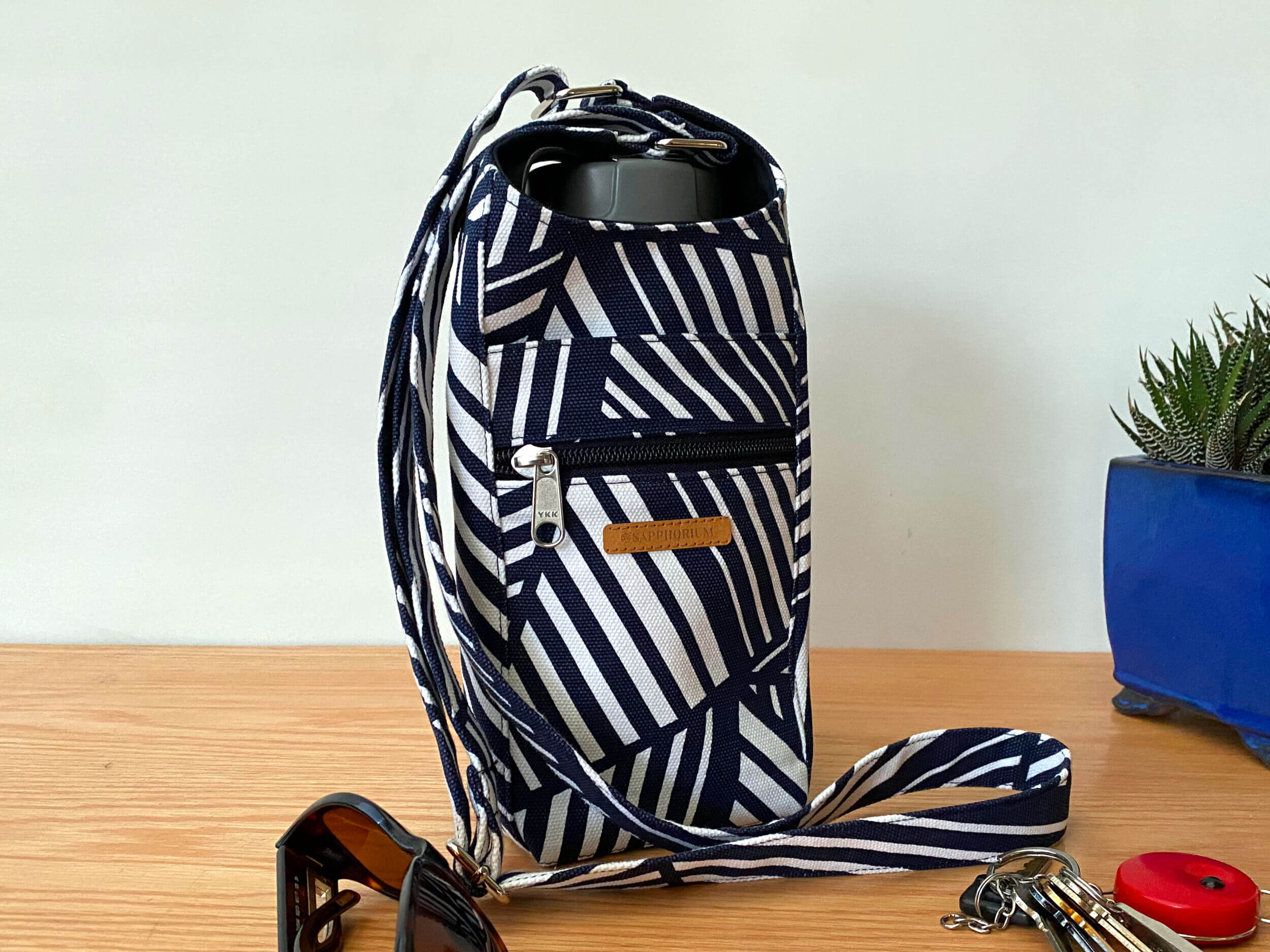 Navy and White striped water bottle bag on a wood grained table with sunglasses, keys with red key ring and plant in a blue pot. Handmade by Sapphorium