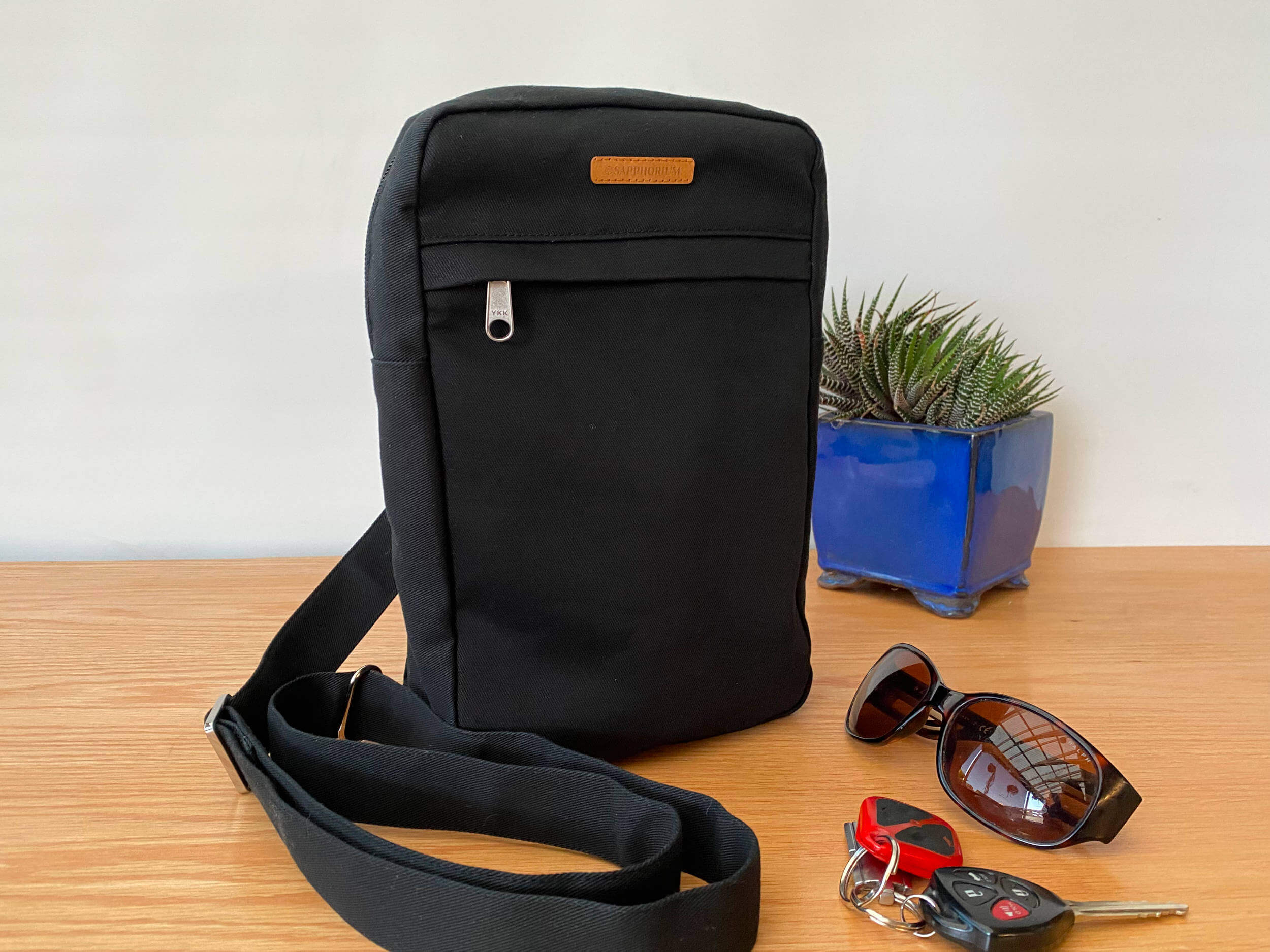 Black Sling Bag on a wooden table next to a green and stripy plant in a shiny blue pot, as well as brown sunglasses and a set of keys. Brown label on bag reads Sapphorium.