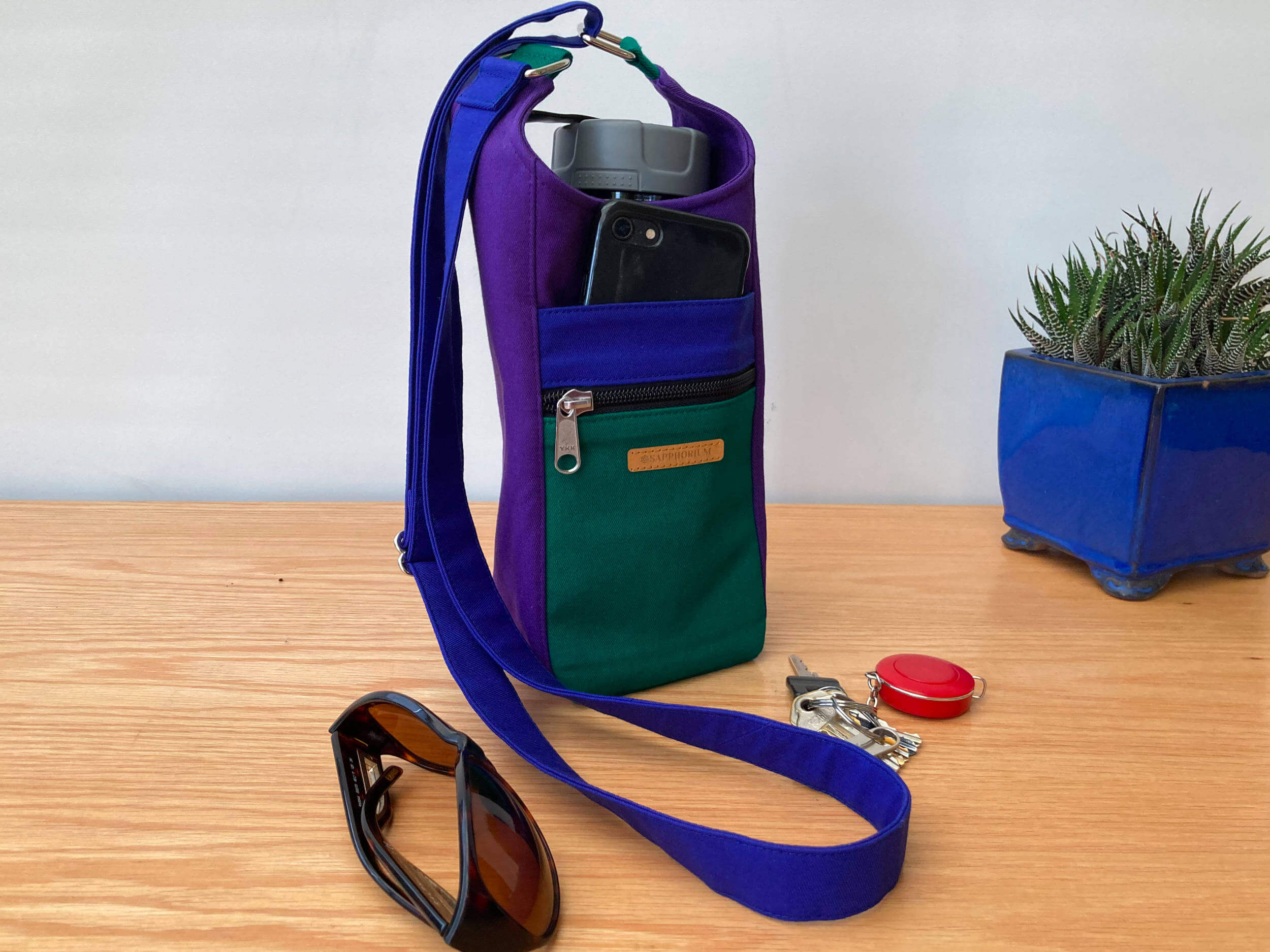 Water Bottle Bag is in Dark Purple, Royal Blue and Jade Green on wooden table. Sunglasses, set of keys and plant in blue pot.