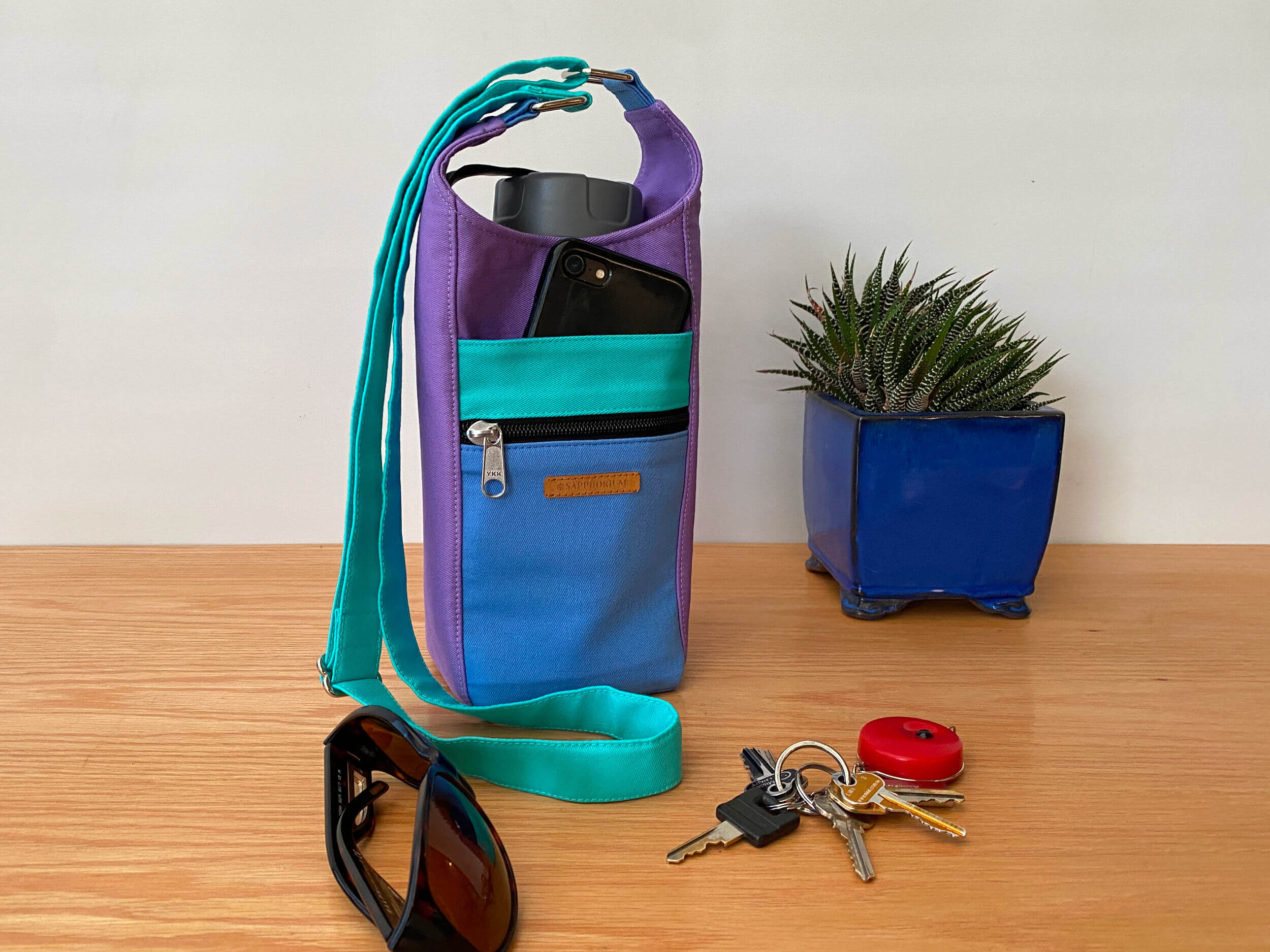 Water Bottle Bag in Light Purple, Periwinkle and Seafoam with a grey lidded water bottle and a black mobile phone in front slip pocket. Next to the bag is a pair of sunglasses, keys with a red key ring and a plant in blue pot. Handmade by Sapphorium.