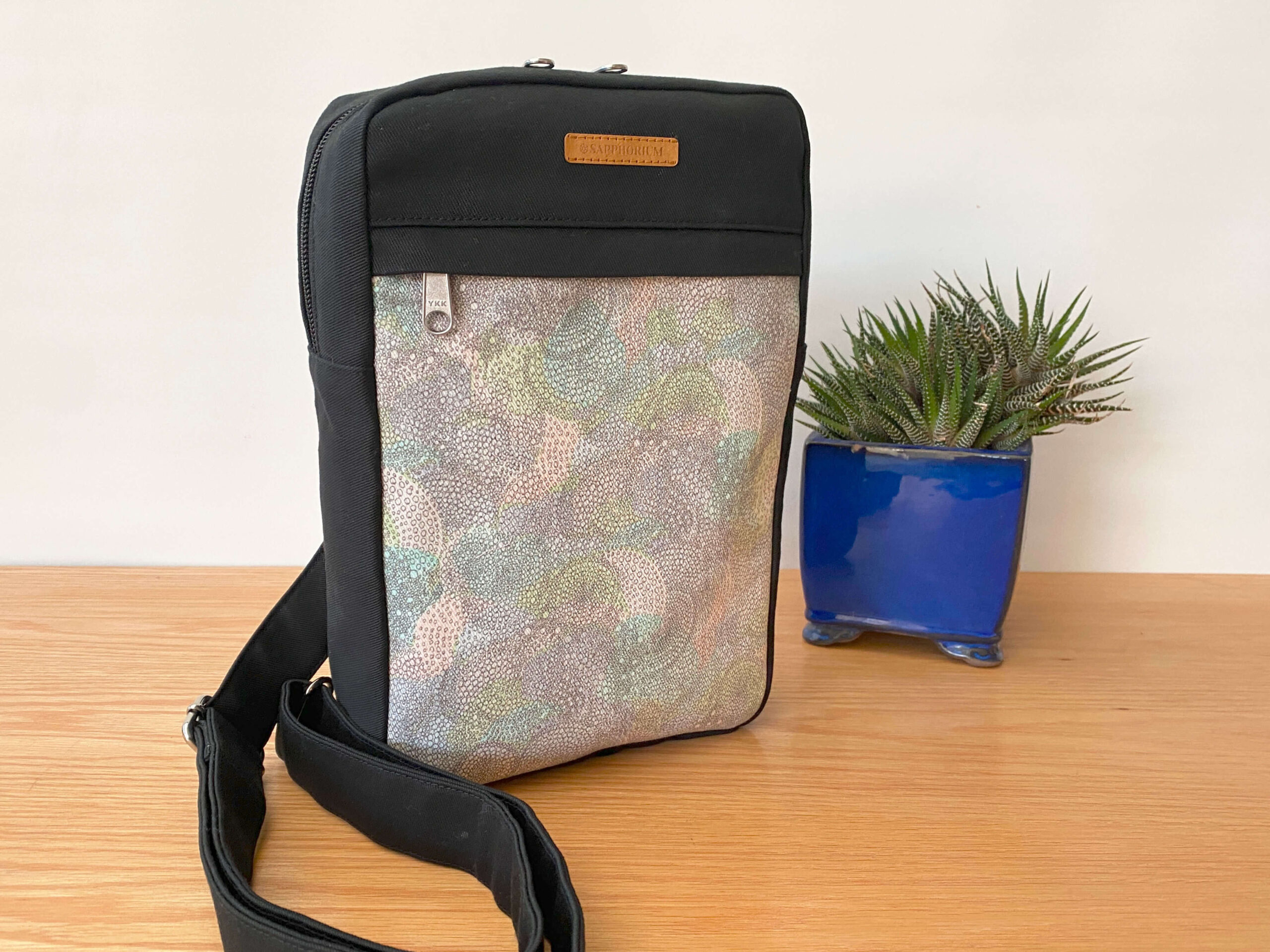 Sling bag in black and grey with pastel swirls on a wooden table, next to a green plant in a blue pot. Handmade by Sapphorium.