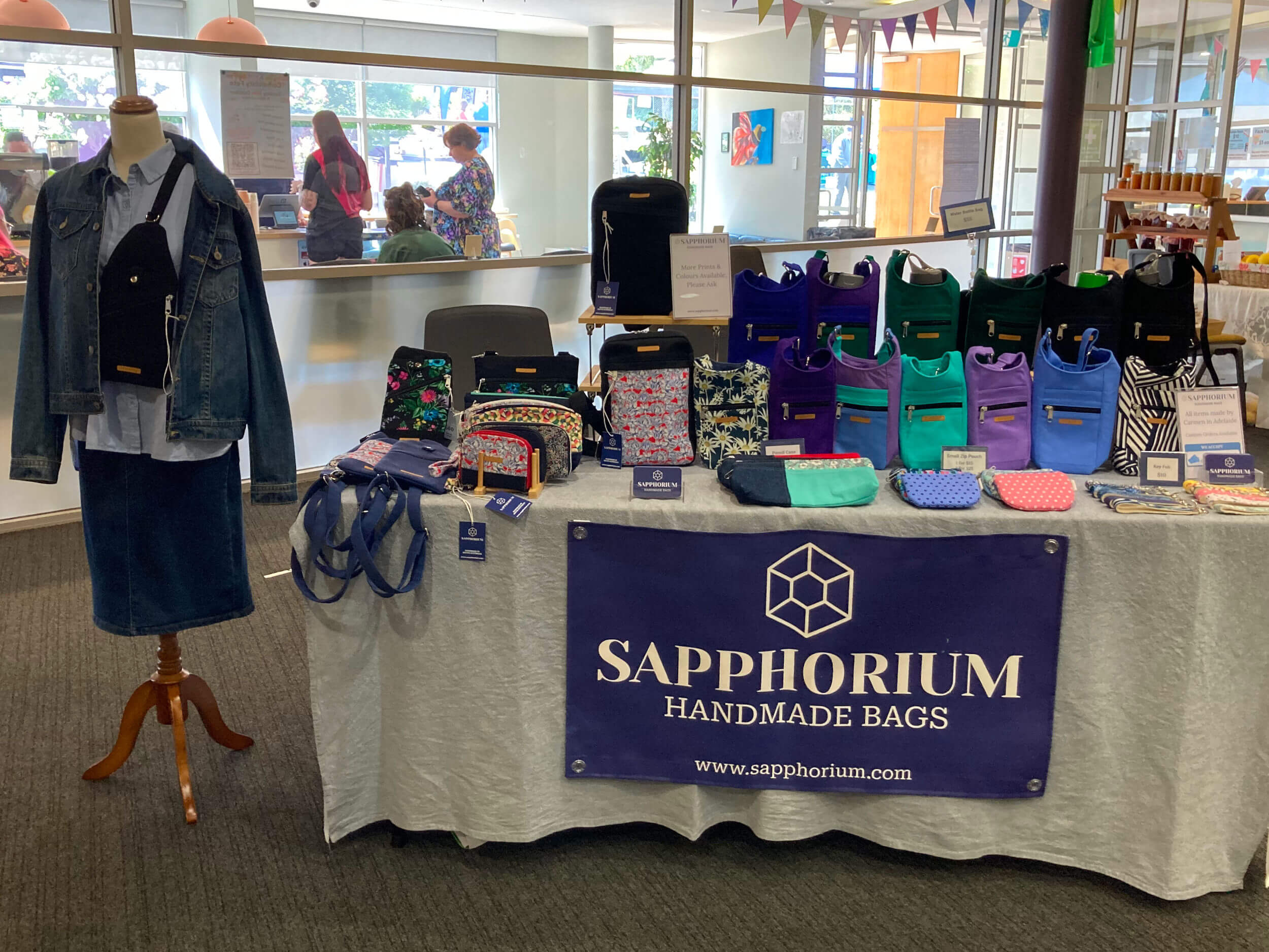 Table at market with bags, wallets and purses on it with large sign reading Sapphorium - Handmade Bags. On the side is a mannequin dressed in denim jacket, blue shirt and skirt with a black bag.