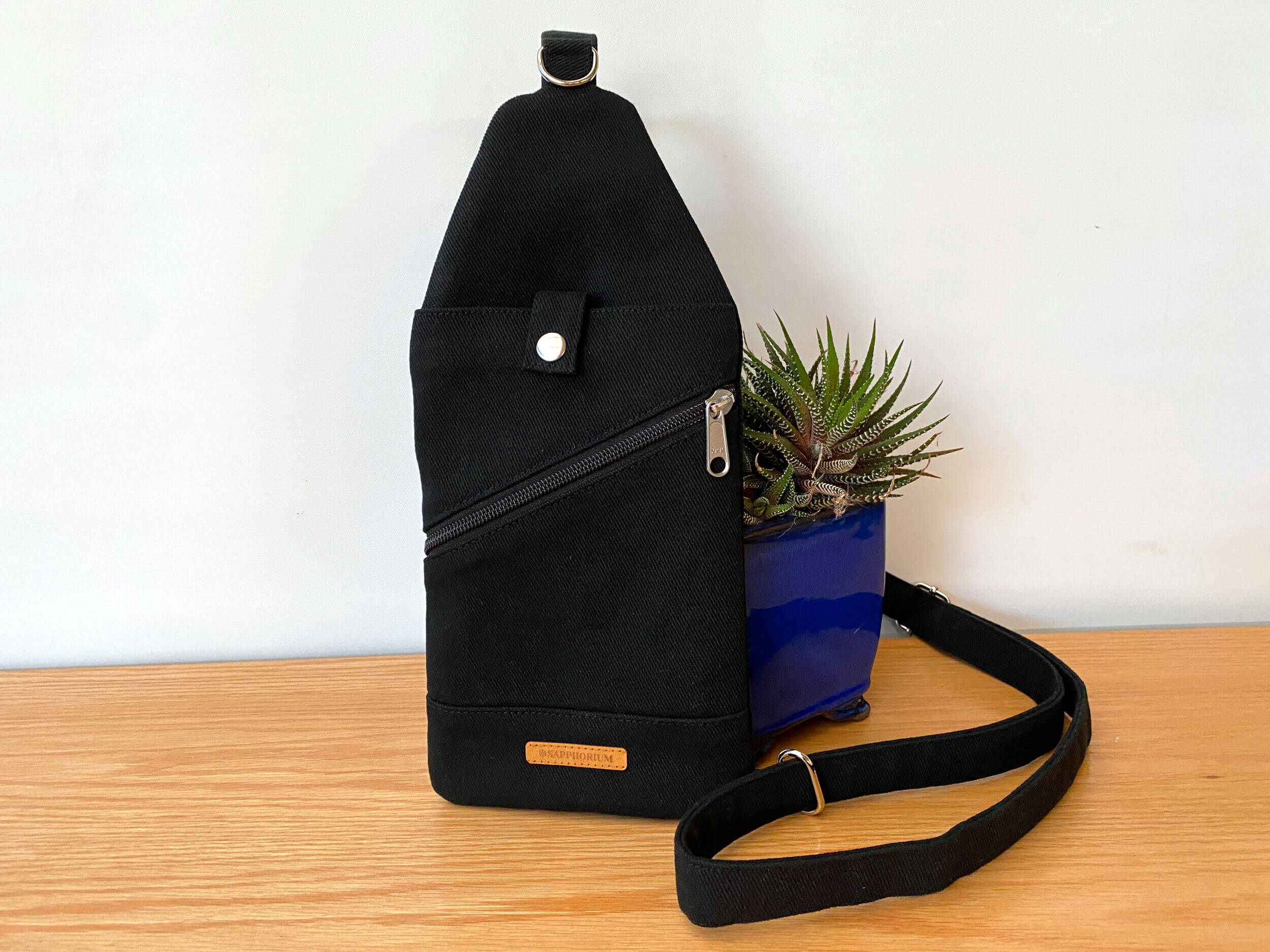 Black bag with snap closure on slip pocket and diagonal zipper pocket. Behind it is a green plant, sitting on a wooden table.