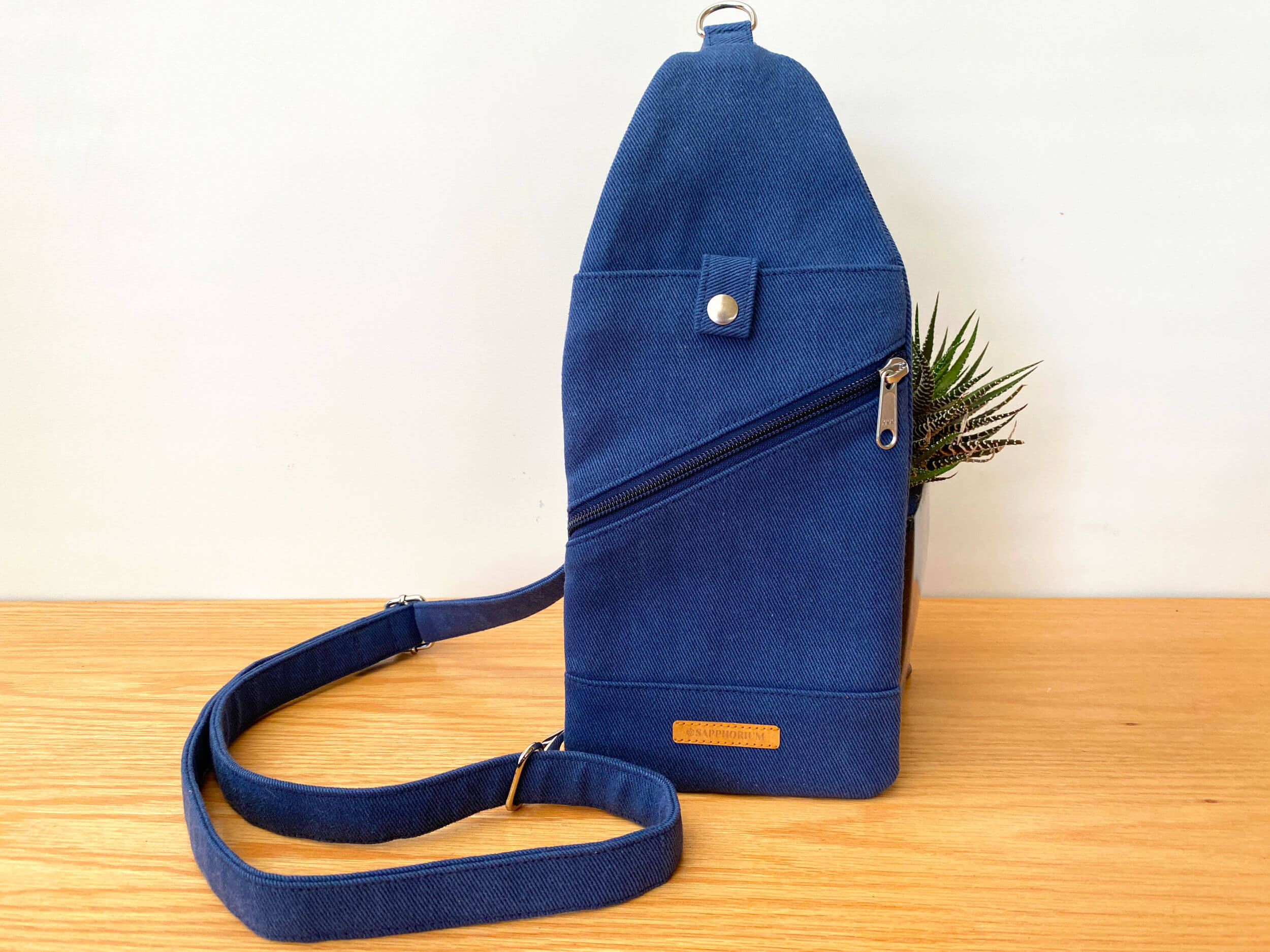 Navy blue sling bag with top pocket with press stud closure, diagonal zipper pocket, on a wooden table.