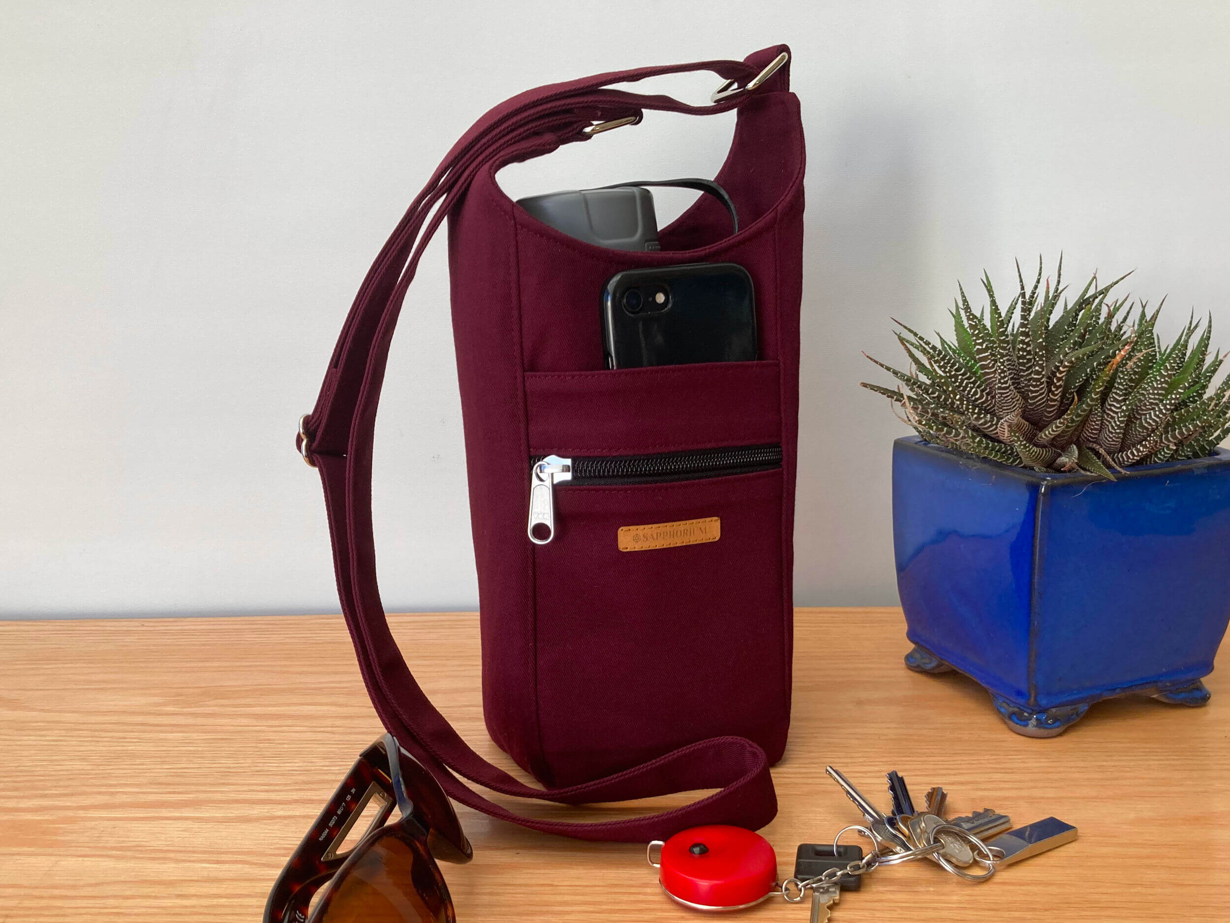 Burgundy Water Bottle Bag with grey lidded water bottle in it, a black phone in the front pocket. To side is a plant in a shiny blue pot. In front is set of keys and sunglasses. On a wooden table. Made by Sapphorium.