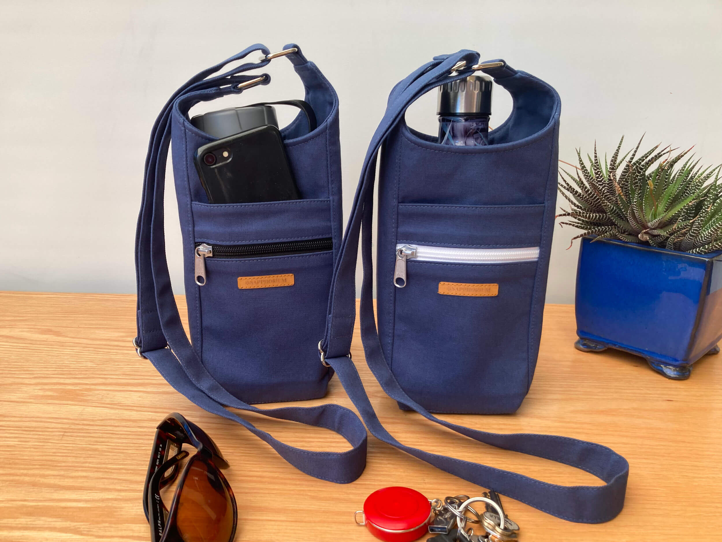 Two Marine Blue Water Bottle Bags one with a black zip and the other with white zip. To side is a plant in a shiny blue pot. In front is set of keys, black phone and sunglasses. On a wooden table. Made by Sapphorium.