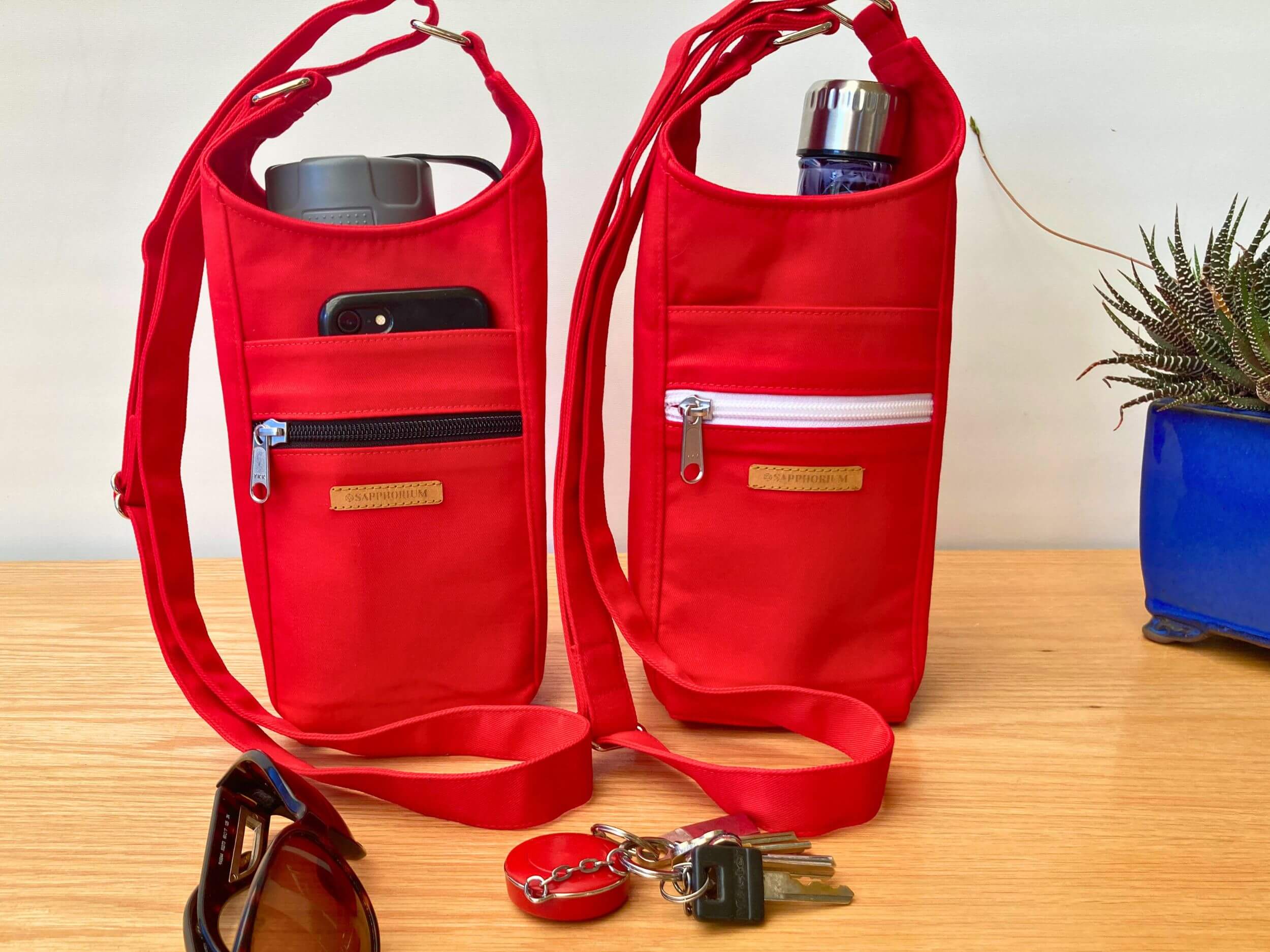 Two Red Blue Water Bottle Bags one with a black zip and the other with white zip. To side is a plant in a shiny blue pot. In front is set of keys and sunglasses. On a wooden table. Made by Sapphorium.