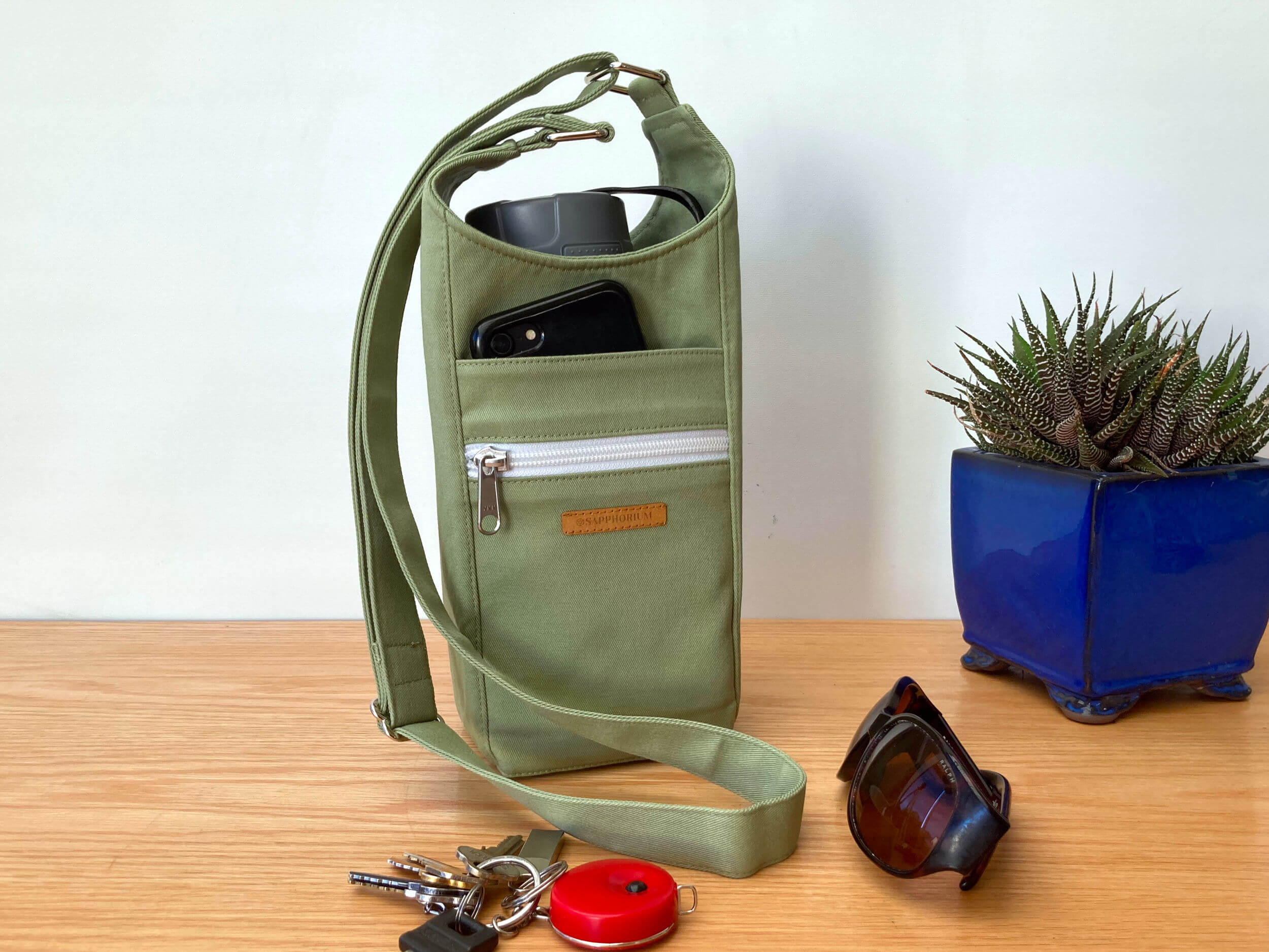 Sage green Water Bottle Bag with grey lidded water bottle in it, a black phone in the front pocket. To side is a plant in a shiny blue pot. In front is set of keys and sunglasses. On a wooden table. Made by Sapphorium.