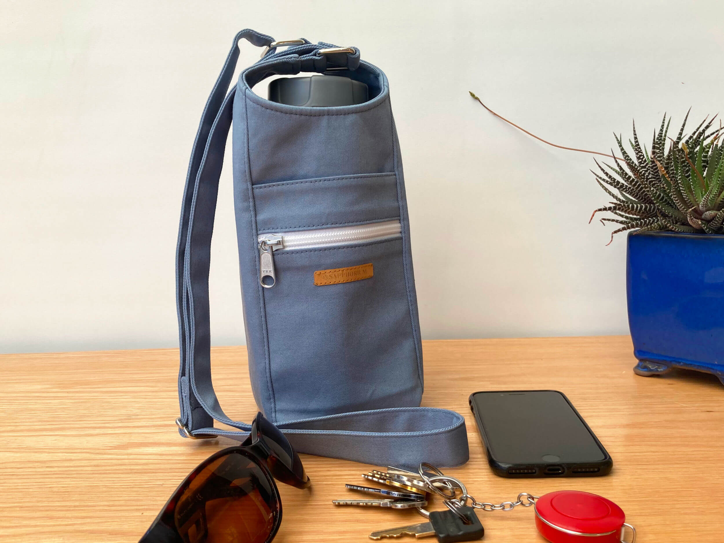 Slate Blue Water Bottle Bag with grey lidded water bottle in it. To side is a plant in a shiny blue pot. In front is set of keys, black phone and sunglasses. On a wooden table. Made by Sapphorium.