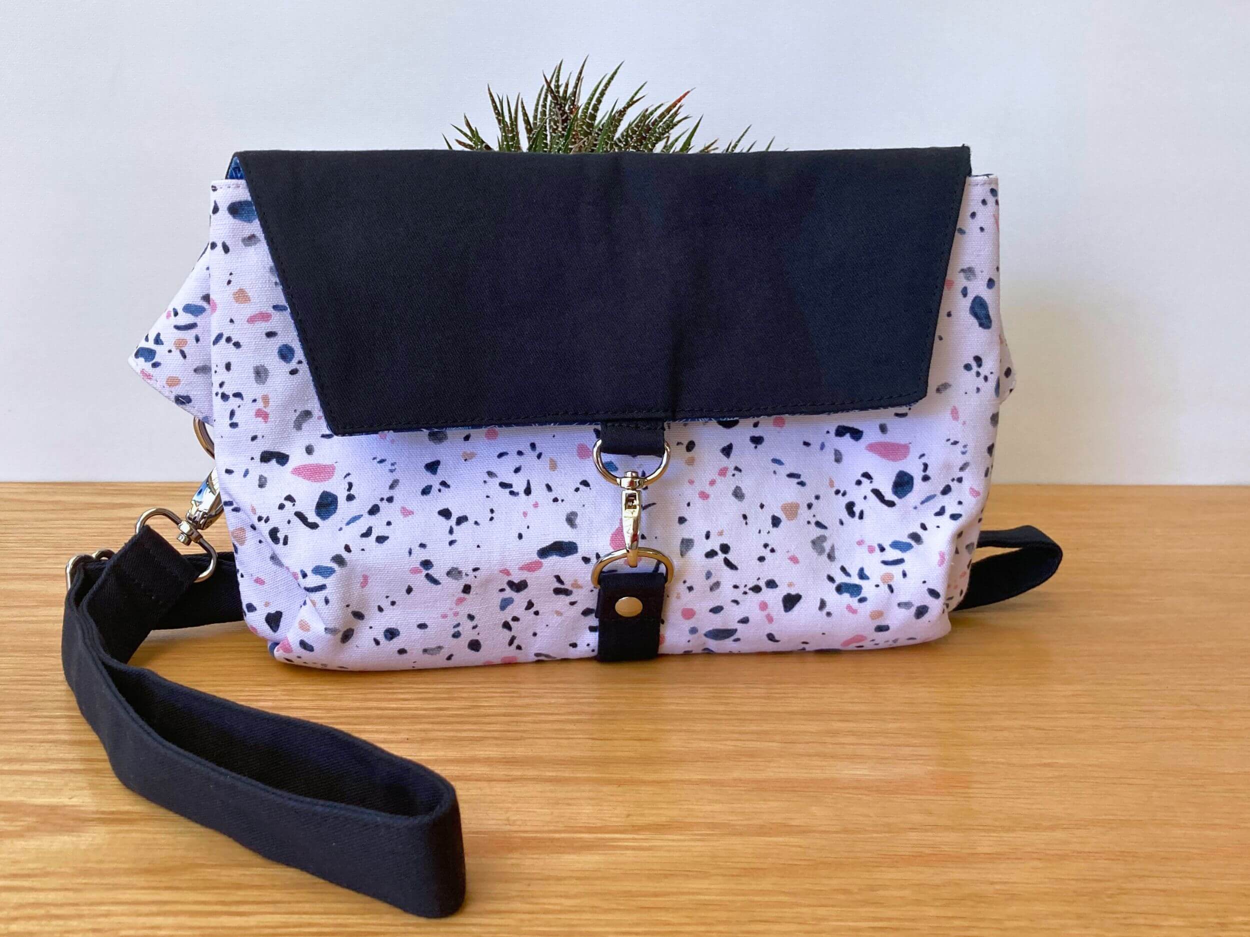 Rectangular bag with navy top flap and White Marble bottom section. Navy strap is folded to one side of the bag and has silver hardware. Behind the bag is green and white fleshy plant. On a wooden table. Handmade by Sapphorium.