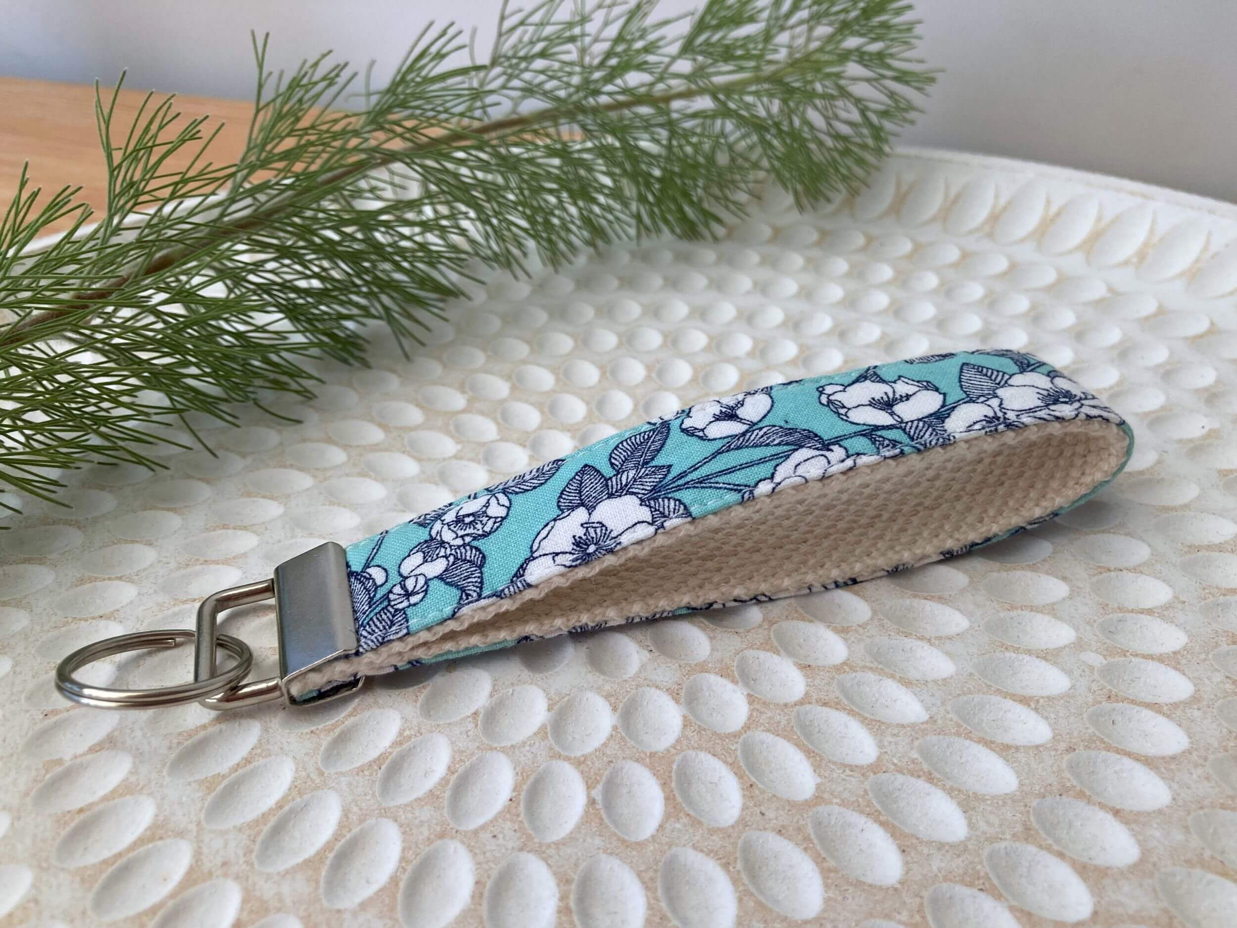 Key fob in an aqua rose print, on a textured white plate with fine leaved plant cutting in top left corner. Handmade by Sapphorium.