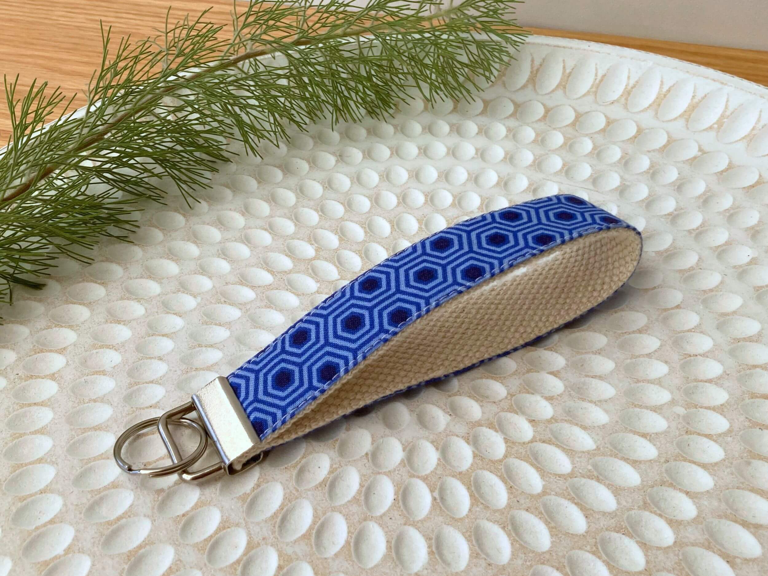 Key fob in a blue hexagon print, on a textured white plate with fine leaved plant cutting in top left corner. Handmade by Sapphorium.