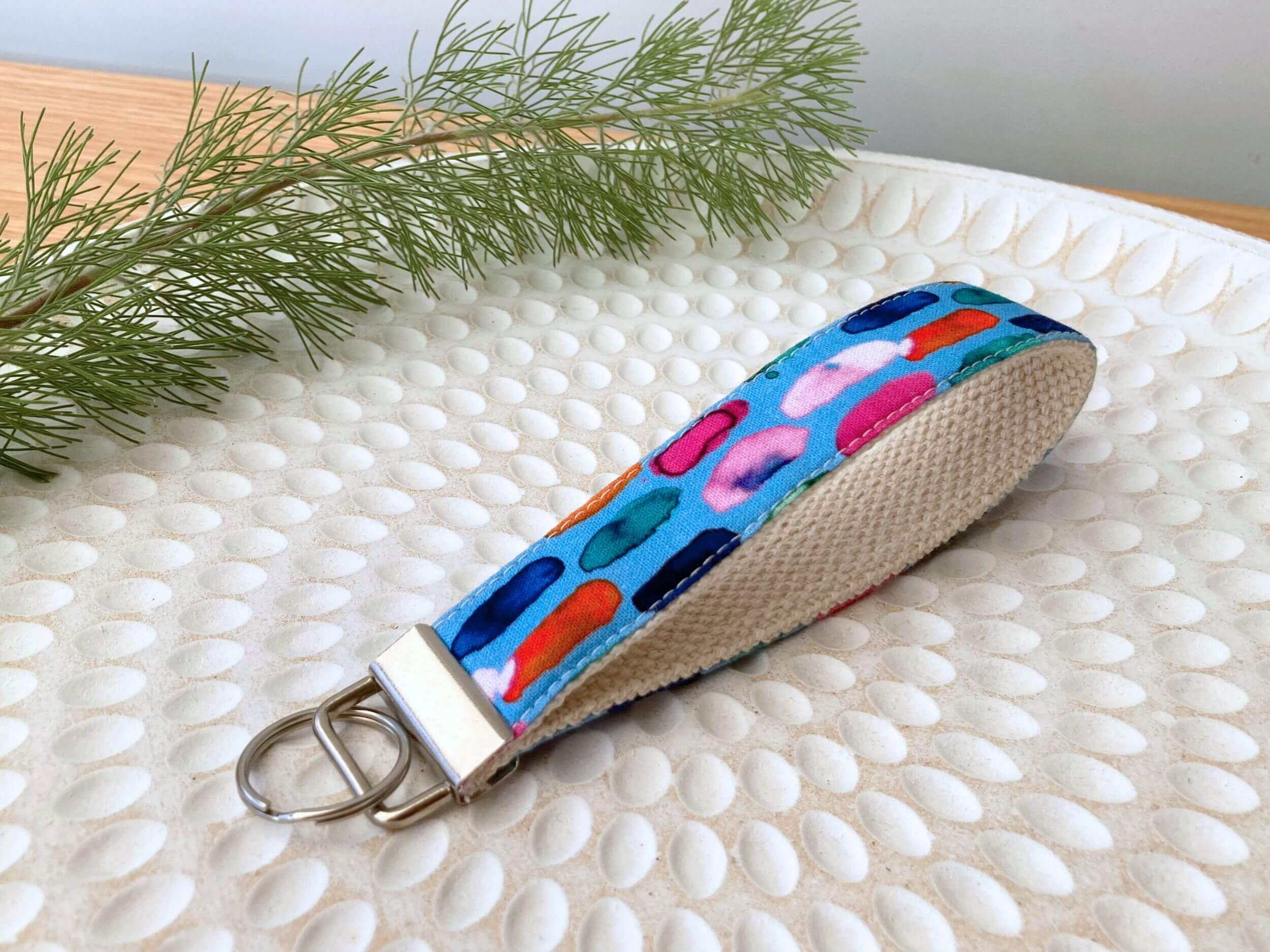 Key fob in a blue with multicolored dashes print, on a textured white plate with fine leaved plant cutting in top left corner. Handmade by Sapphorium.