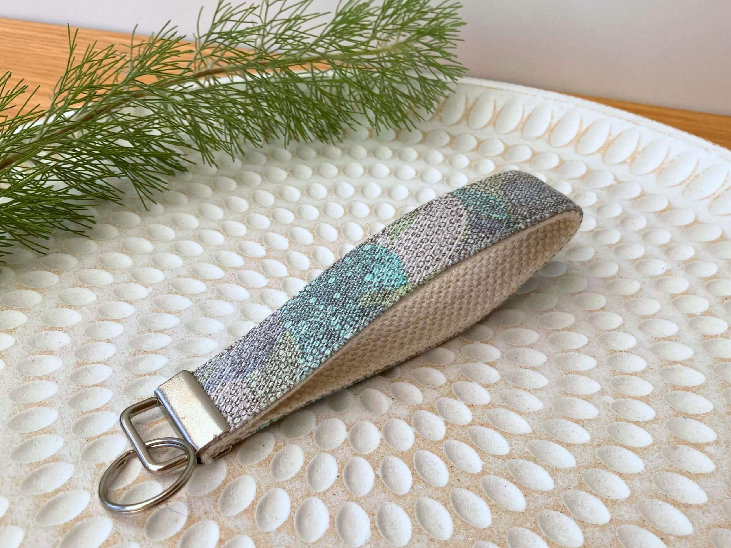 Key fob in a grey and pastels Liberty print, on a textured white plate with fine leaved plant cutting in top left corner. Handmade by Sapphorium.