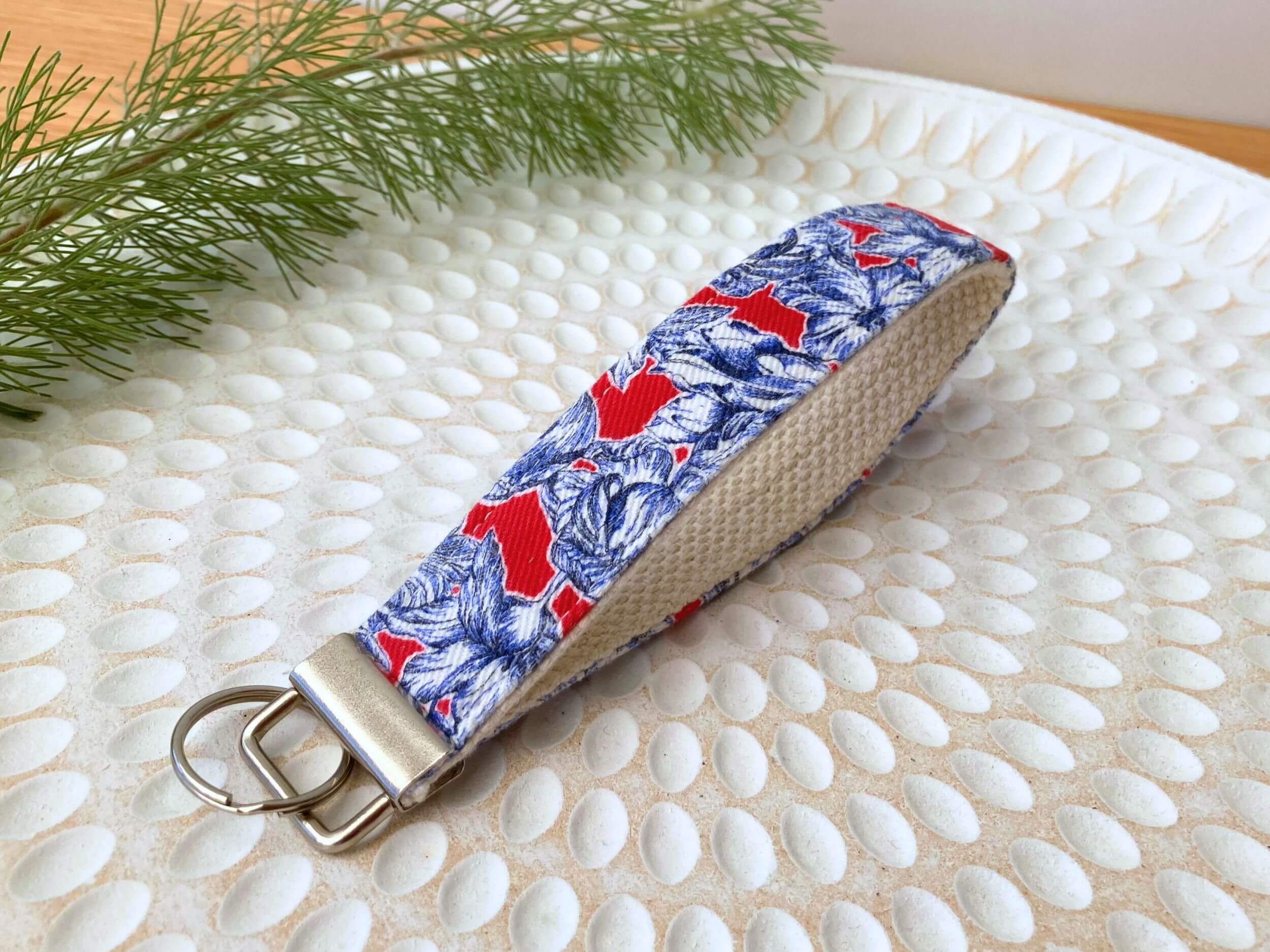 Key fob in a blue floral on red print, on a textured white plate with fine leaved plant cutting in top left corner. Handmade by Sapphorium.