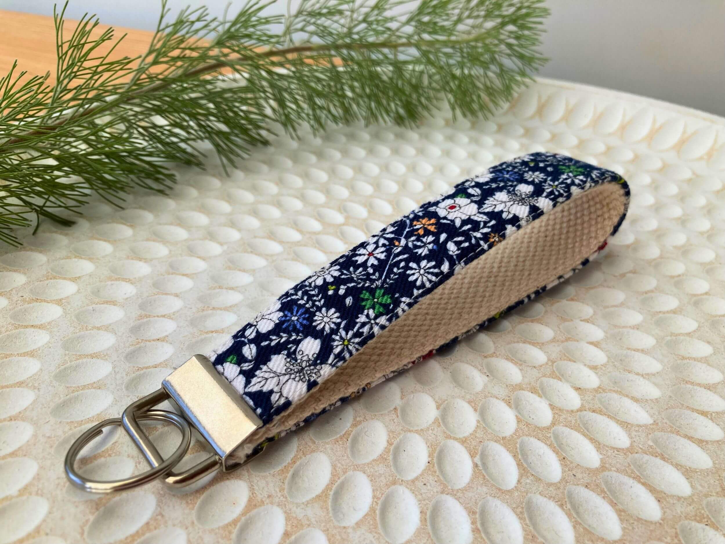 Key fob in a white daisies on navy print, on a textured white plate with fine leaved plant cutting in top left corner. Handmade by Sapphorium.