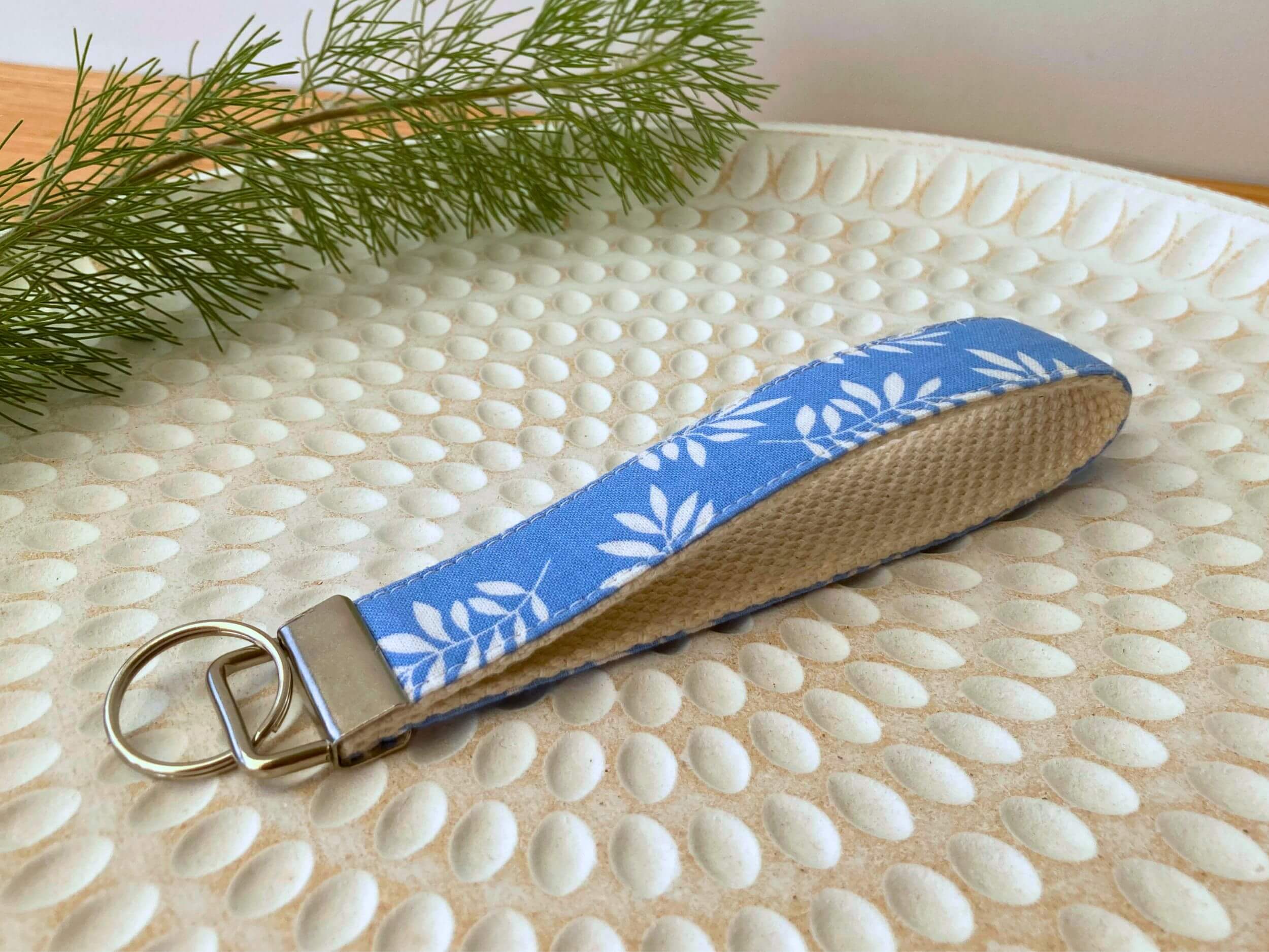 Key fob in a white leaves on light blue print, on a textured white plate with fine leaved plant cutting in top left corner. Handmade by Sapphorium.