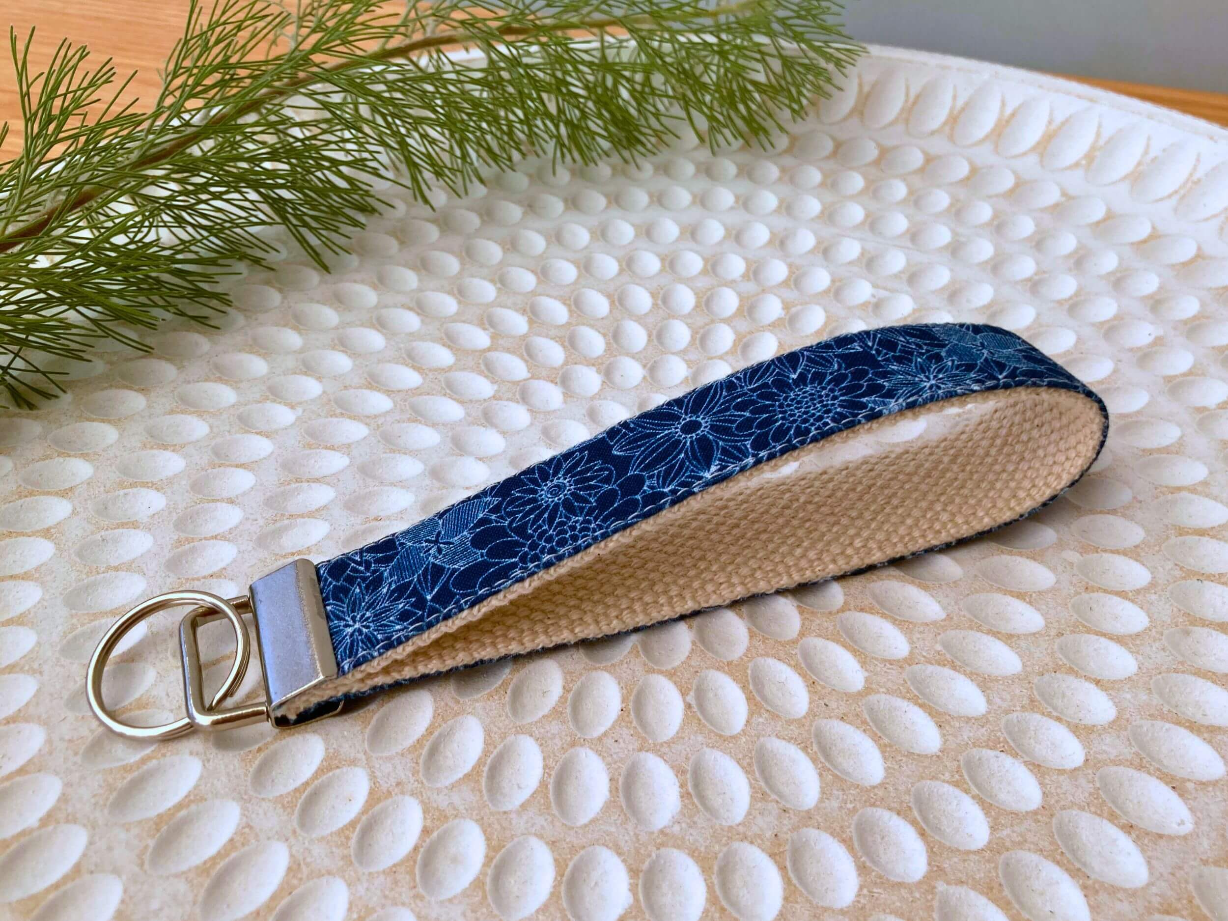 Three key fobs in navy floral print, on a textured white plate with fine leaved plant cutting in top left corner. Handmade by Sapphorium.