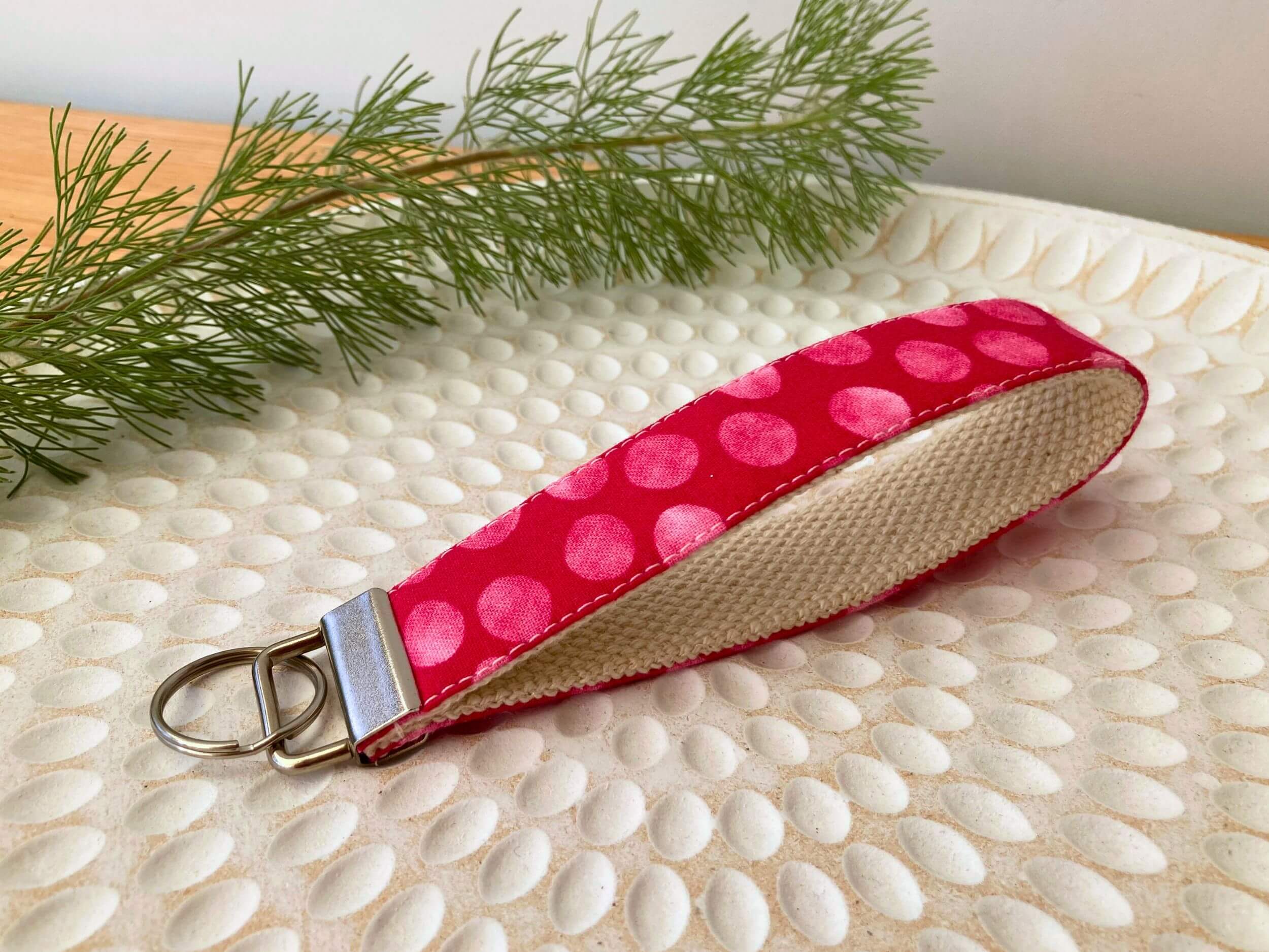 Key fob in a pink with spots print, on a textured white plate with fine leaved plant cutting in top left corner. Handmade by Sapphorium.