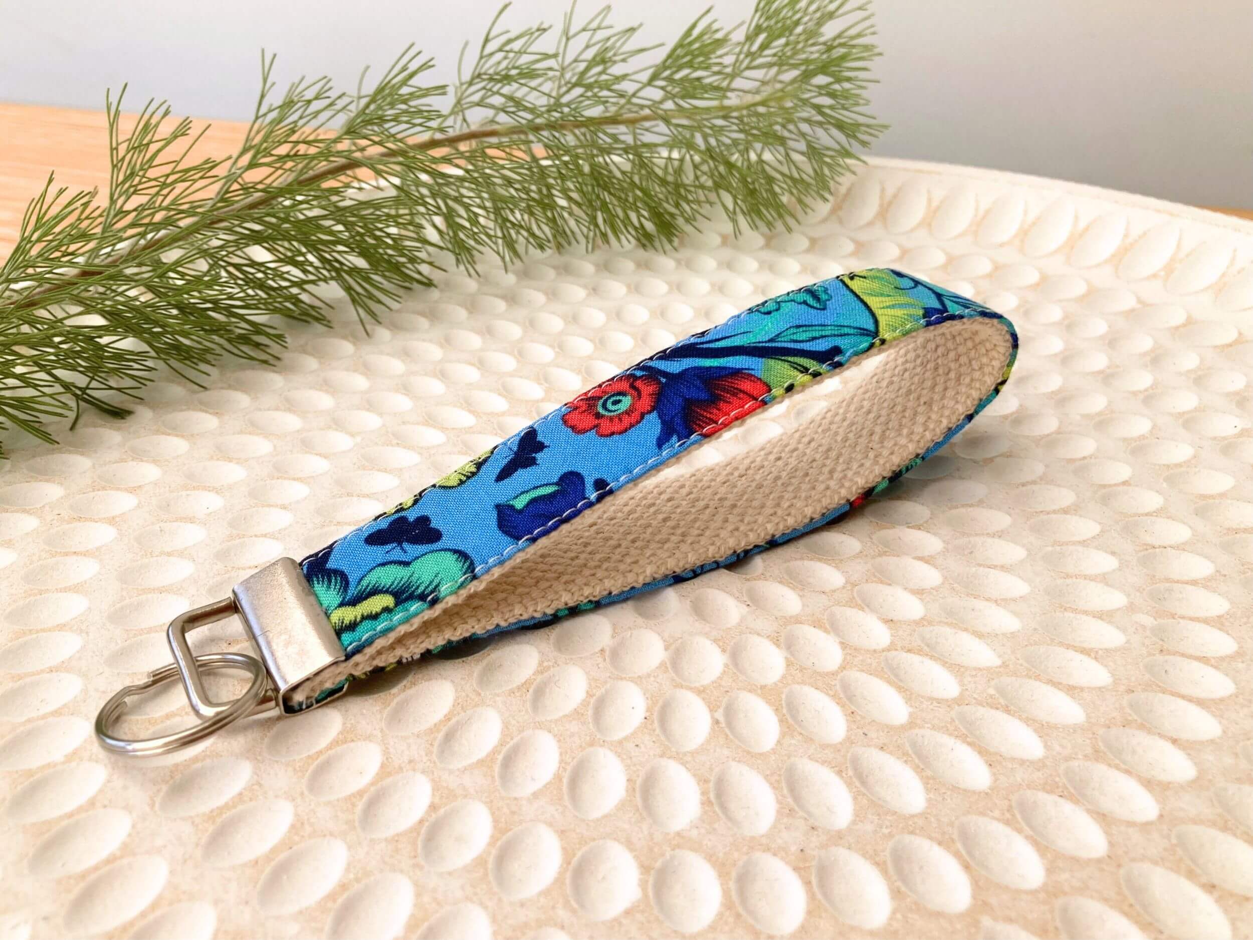 Key fob in a blue tail feathers print, on a textured white plate with fine leaved plant cutting in top left corner. Handmade by Sapphorium.