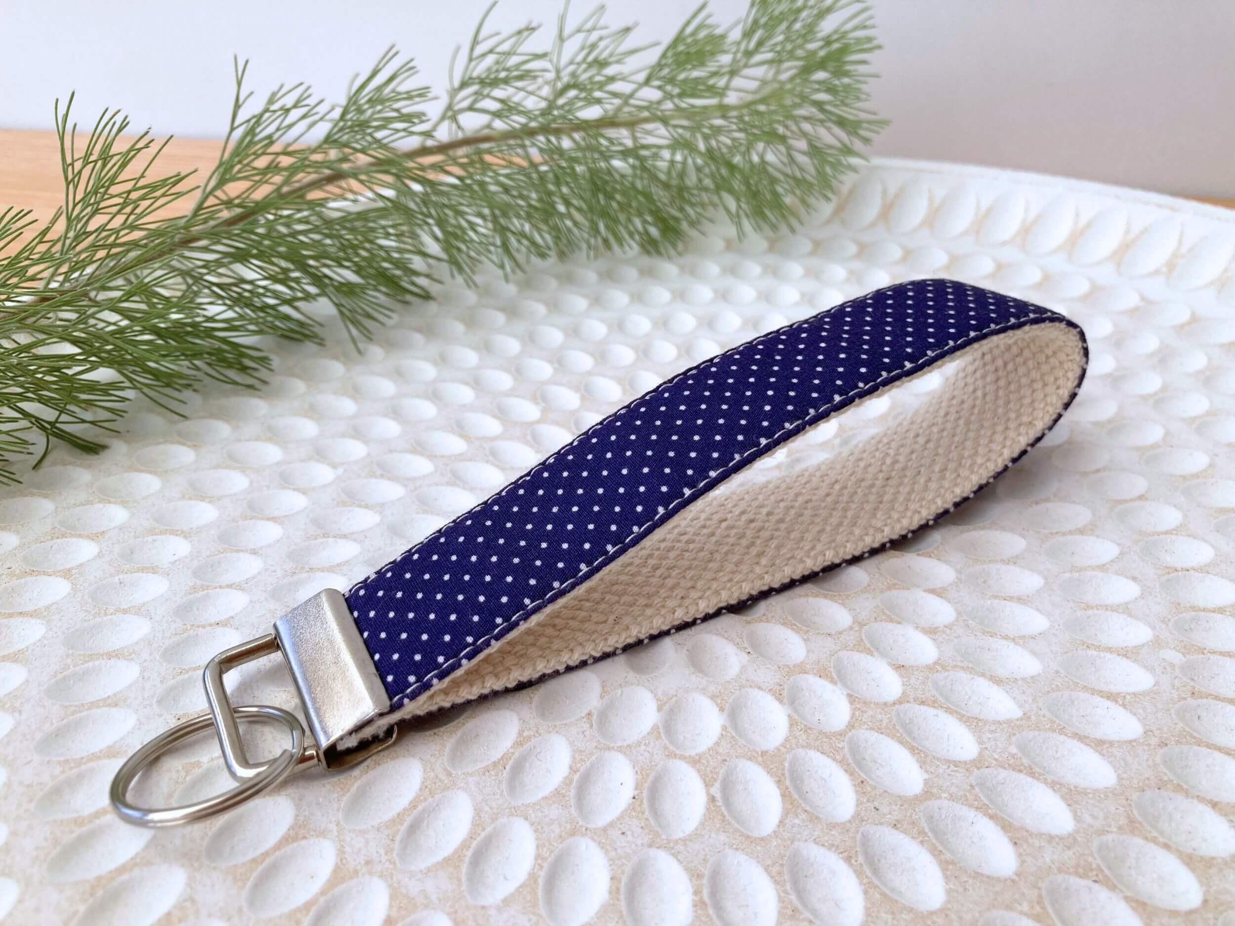Key fob in a navy with small white dots, on a textured white plate with fine leaved plant cutting in top left corner. Handmade by Sapphorium.