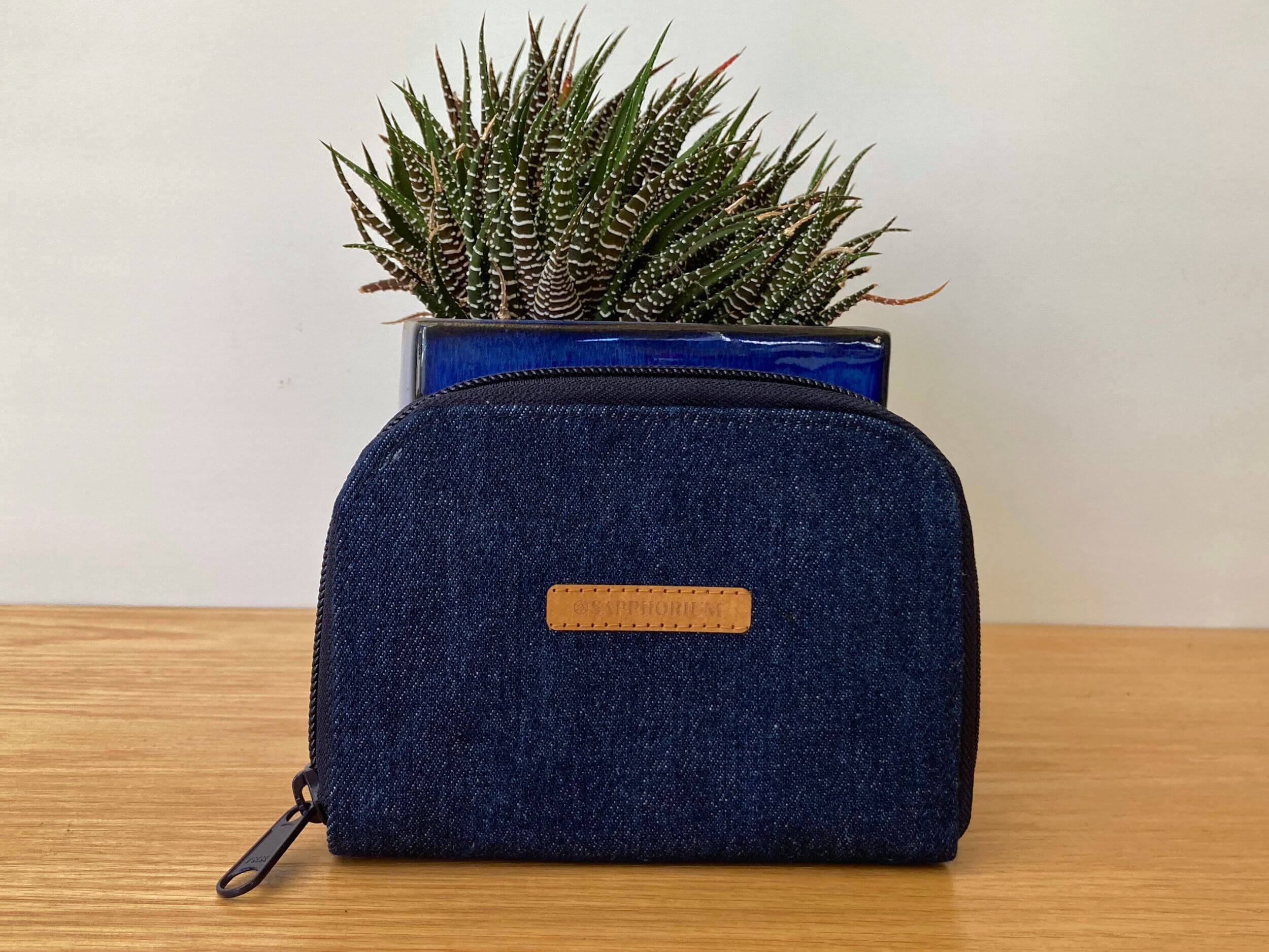 Denim blue zip wallet with brown label with words Sapphorium in front of a shiny blue pot plant with a green and white fleshy leaved plant. On a wooden table. Handmade by Sapphorium.