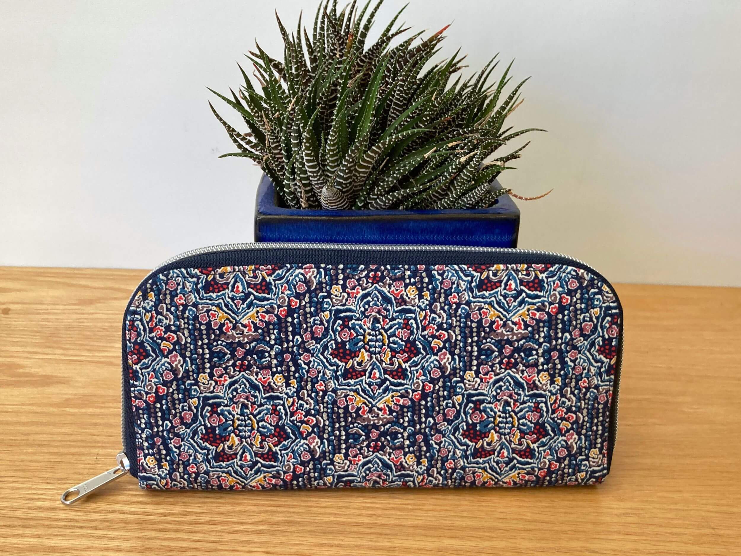 Zip around wallet in a blue and multicoloured repeating pattern with blue and silver zip in front of a shiny blue pot plant with a green and white fleshy leaved plant. On a wooden table. Handmade by Sapphorium.