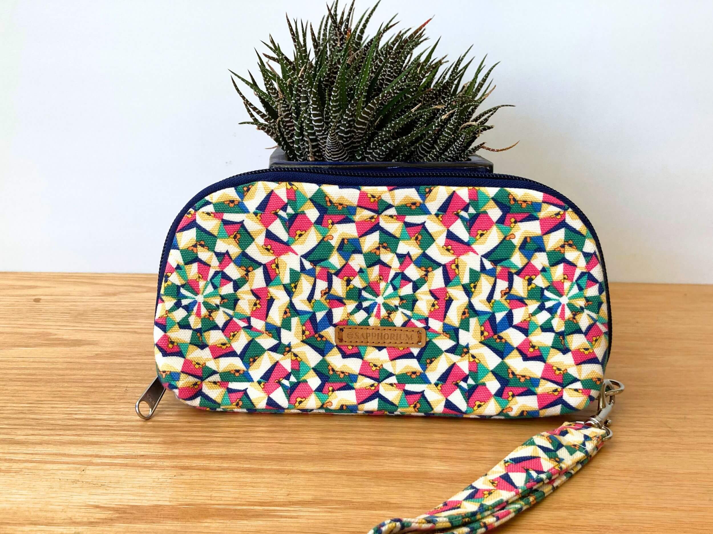 Yellow geometric with blue zip wallet with brown label with words Sapphorium in front of a shiny blue pot plant with a green and white fleshy leaved plant. On a wooden table. Handmade by Sapphorium.