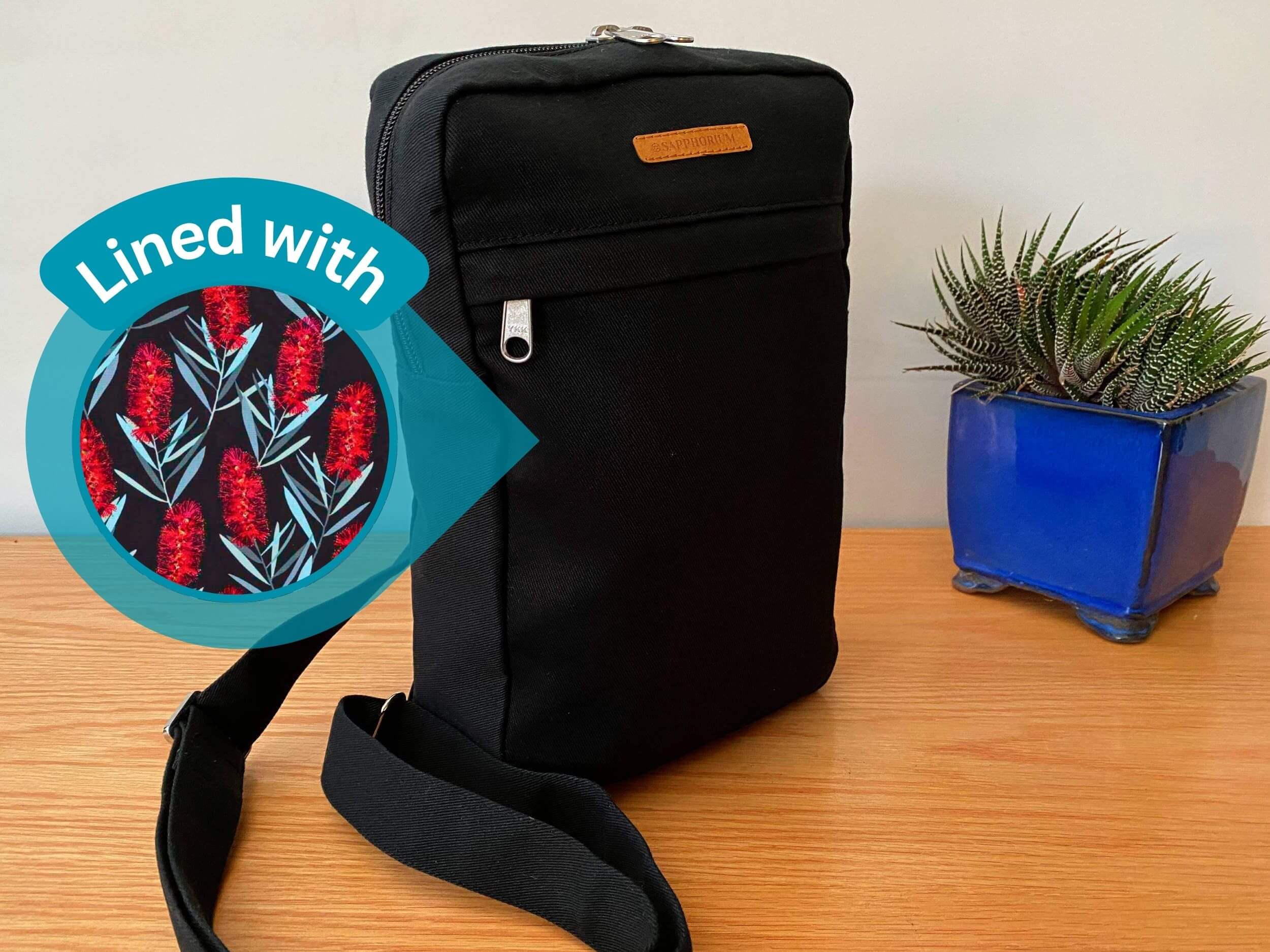 Black Sling Bag on a wooden table next to a green and stripy plant in a shiny blue pot. Brown label on bag reads Sapphorium. Circle on left with text "Lined with" and image of red callistemon on black fabric.