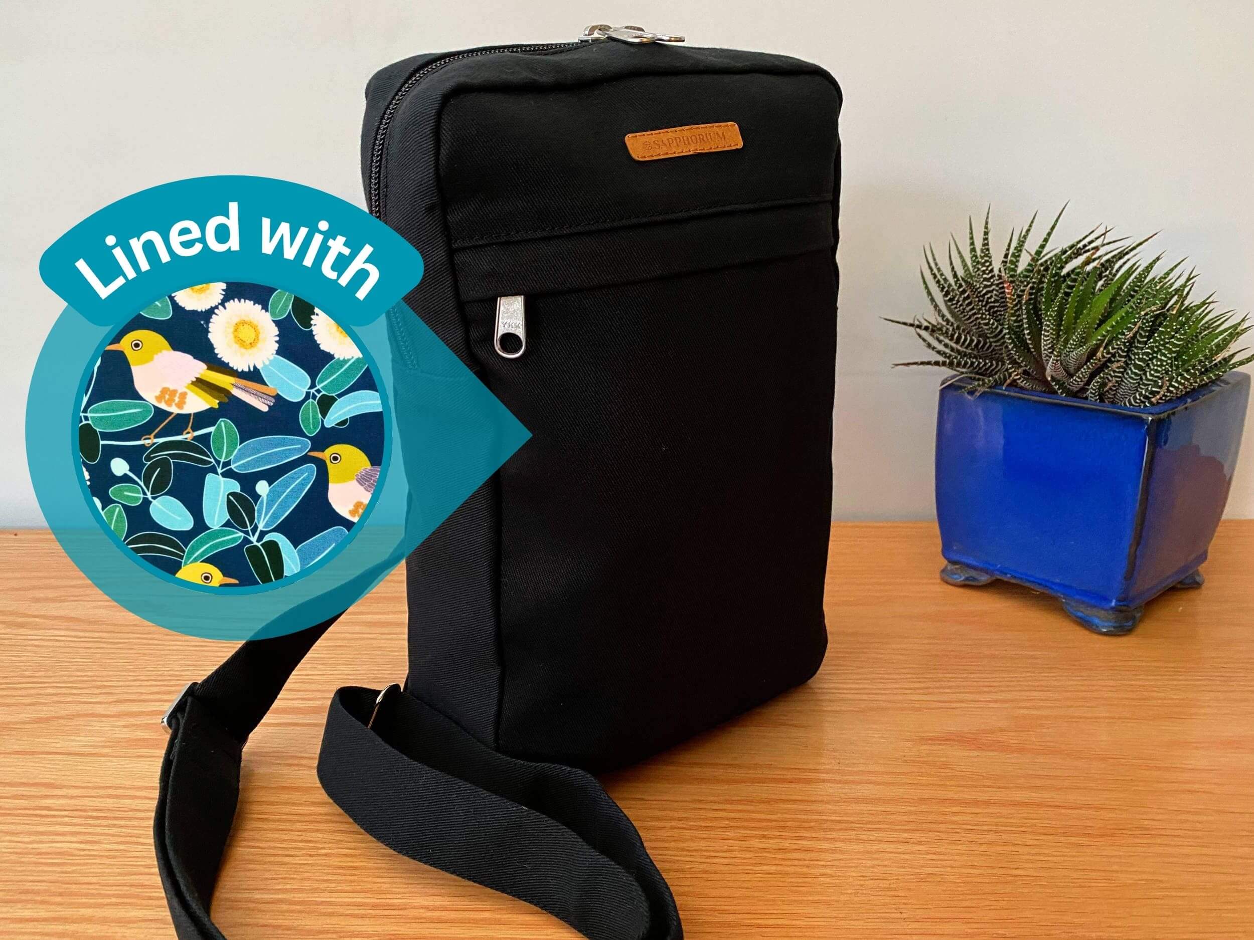 Black Sling Bag on a wooden table next to a green and stripy plant in a shiny blue pot. Brown label on bag reads Sapphorium. Circle on left with text "Lined with" and image of fabric print with bird in mustard, brown and yellow, along with white flowers, green and black leaves, on a teal background .