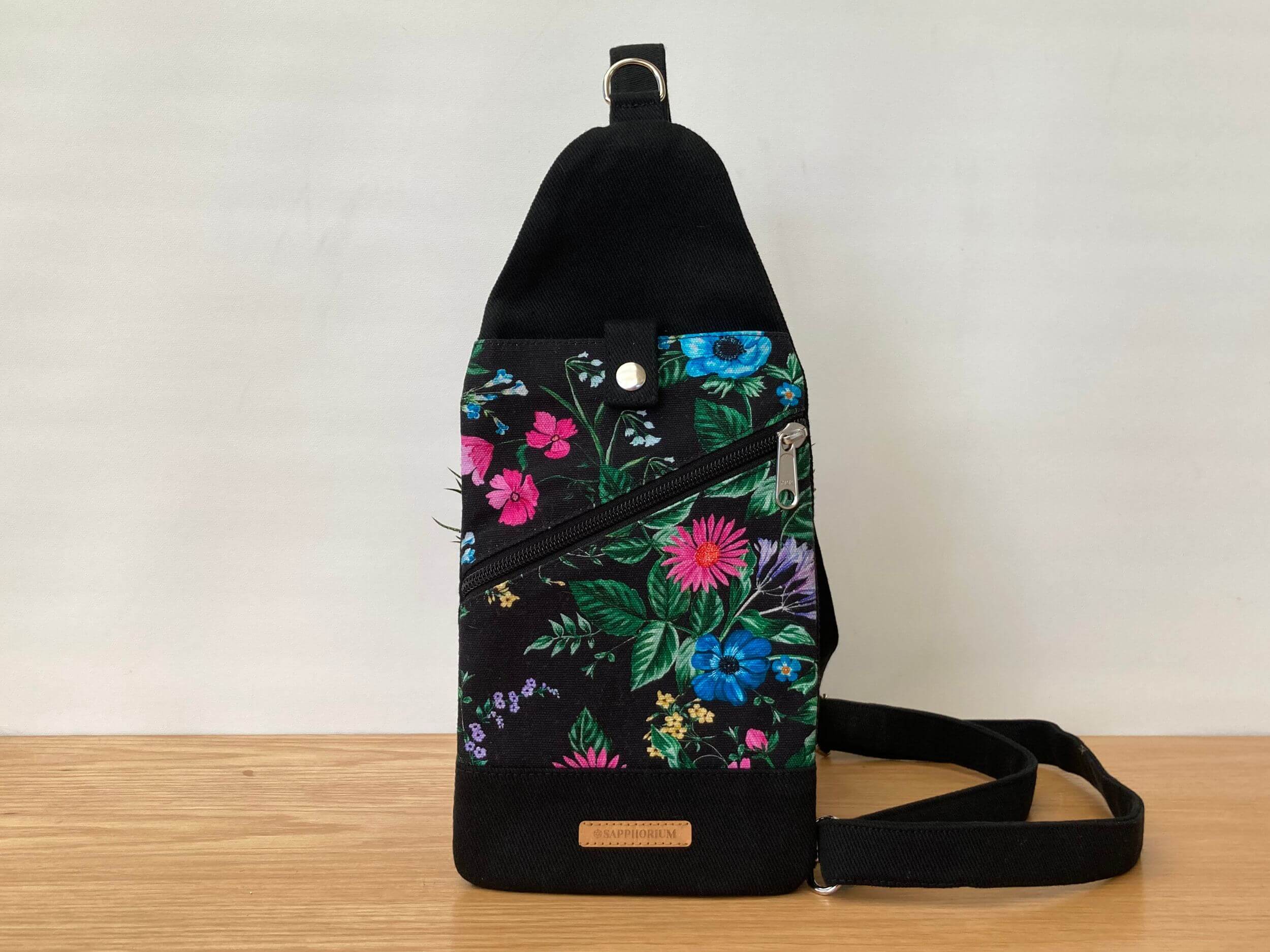 Black and Black floral fabric sling bag with top pocket and press stud closure, diagonal zipper pocket. Leaning against a pot plant, on a wooden table. Handmade by Sapphorium.