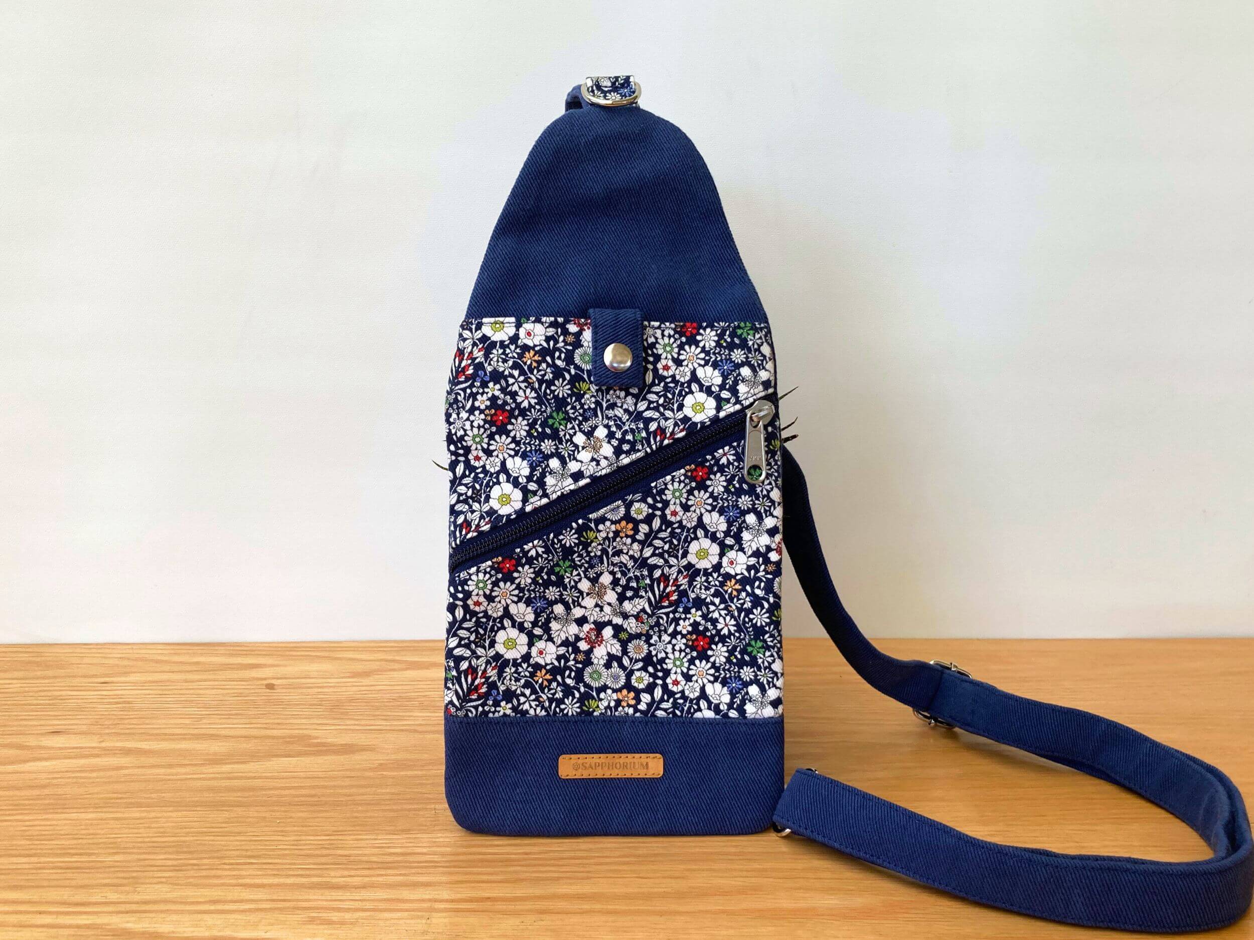 Blue wiht White Daisies sling bag with top pocket and press stud closure, diagonal zipper pocket. Leaning against a pot plant, on a wooden table. Handmade by Sapphorium.