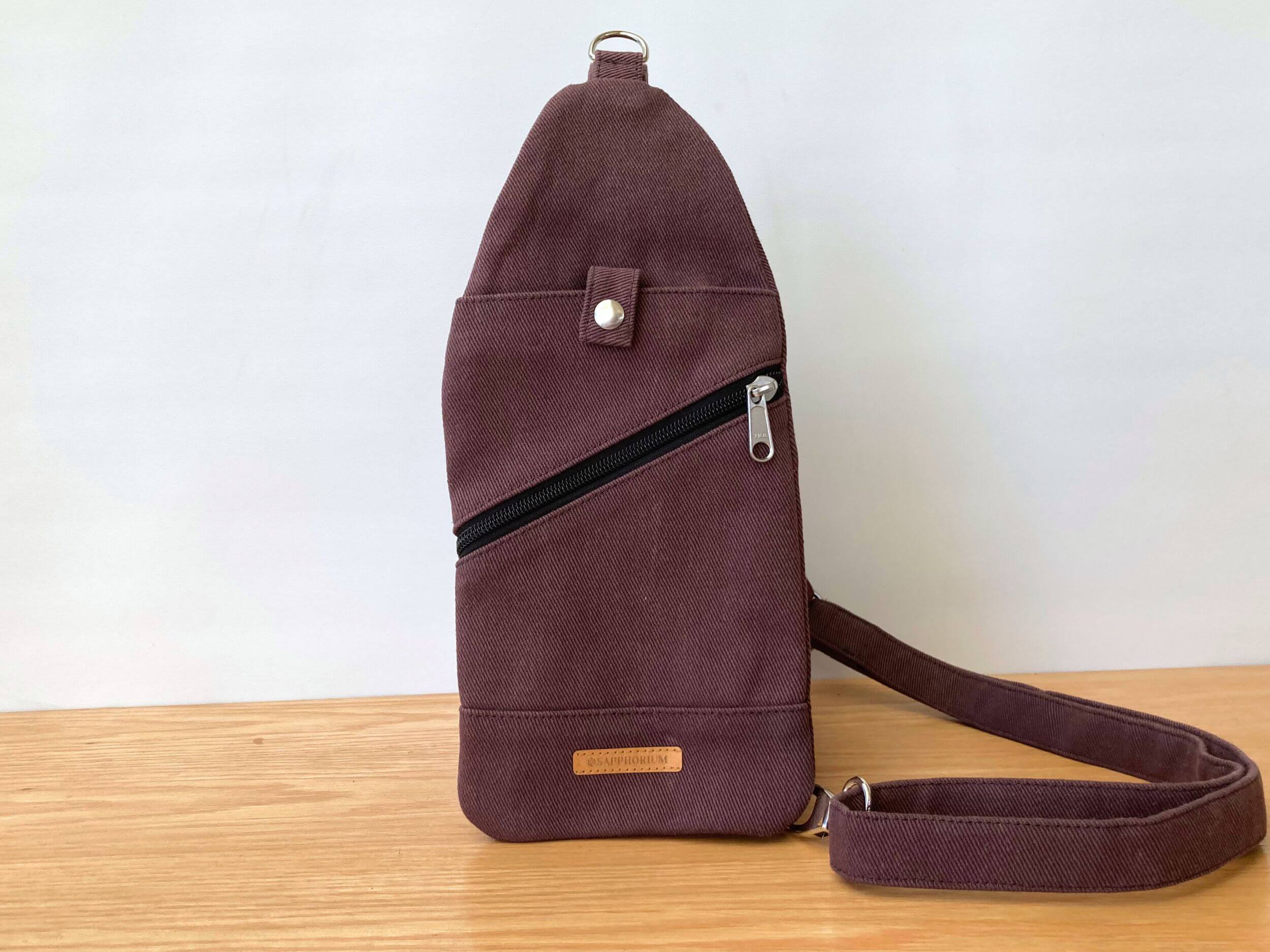 Brown sling bag with top pocket and press stud closure, diagonal black zipper pocket. Leaning against a pot plant, on a wooden table. Handmade by Sapphorium.