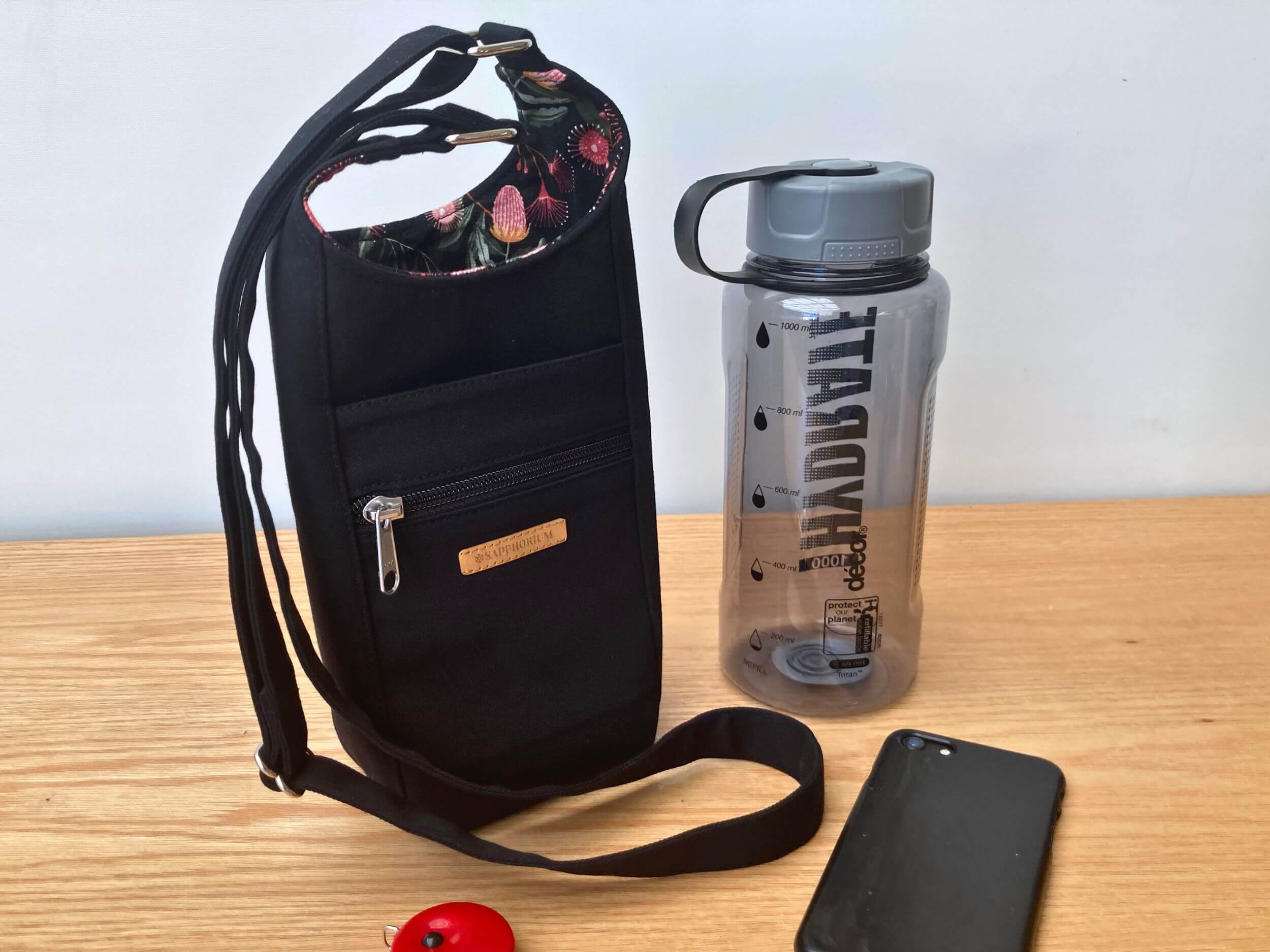 Black with Eucalyptus lining Water Bottle Bag with grey lidded water bottle next to in it. In front is a black phone and red disc for keys. On a wooden table. Made by Sapphorium.